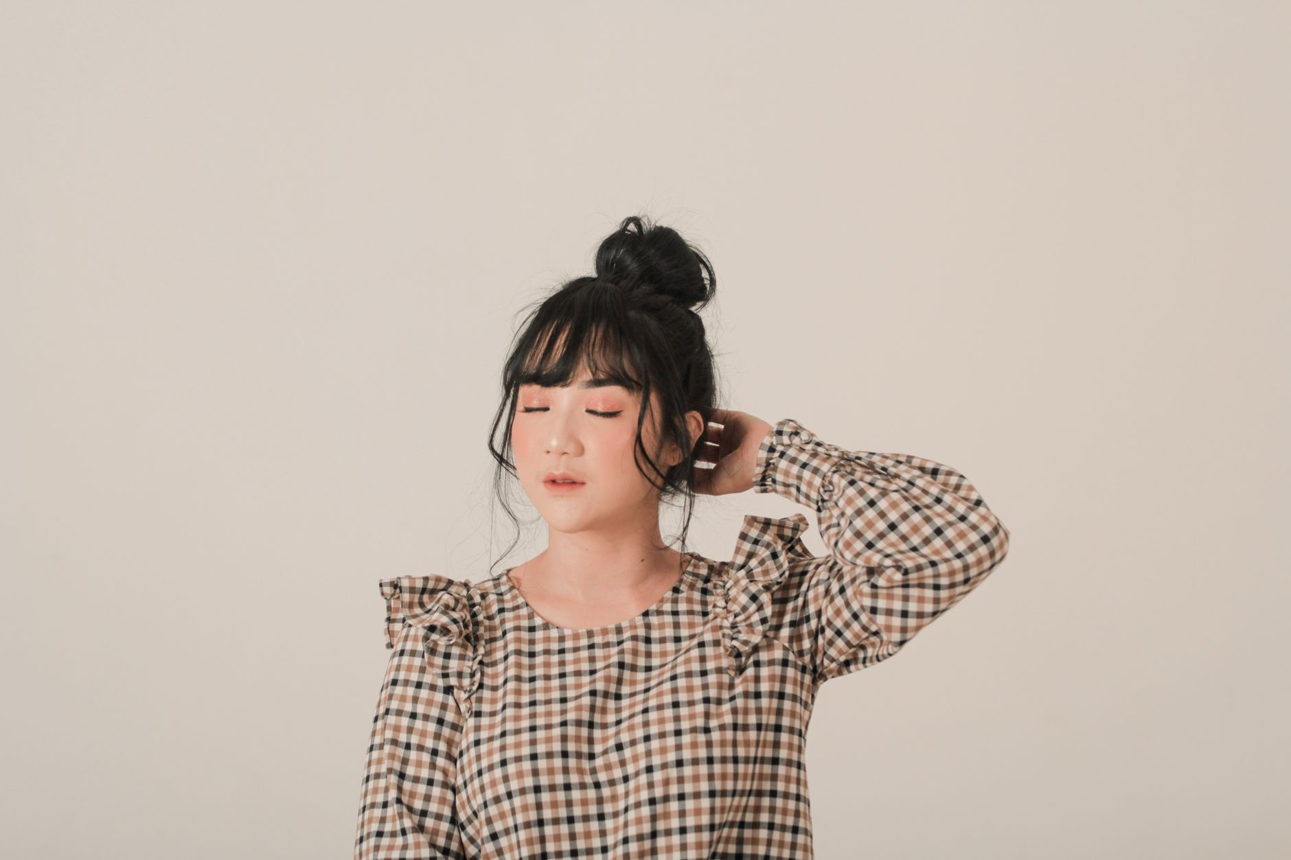 A stylish Asian woman with a bun hairstyle and checked blouse posing in a studio setting.