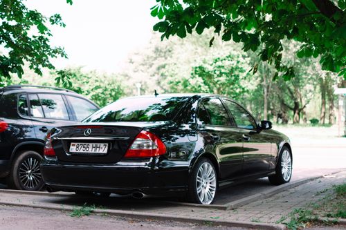 Free A sleek black car parked in an outdoor lot under lush green trees on a sunny day. Stock Photo