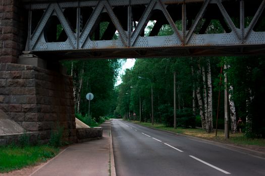A quiet road enveloped by lush greenery, passing under a sturdy steel bridge.