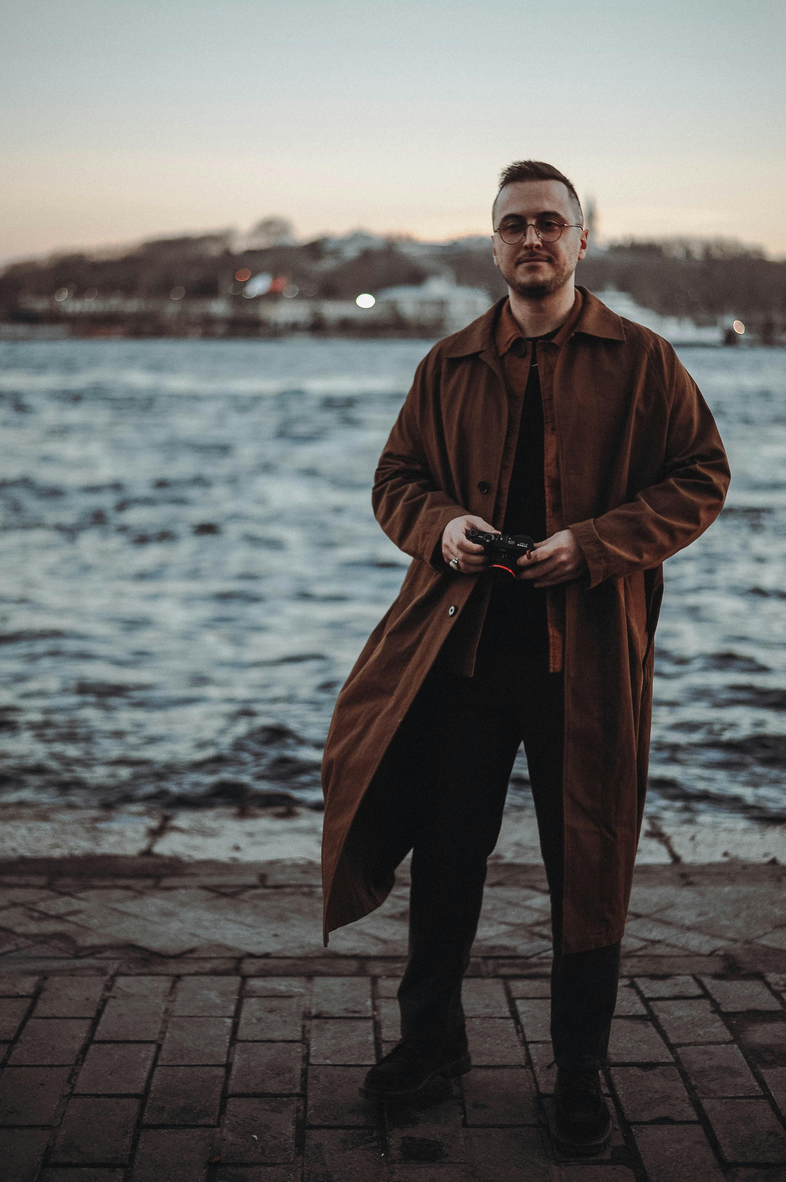 Fashionable man in a brown coat standing by the water during twilight, exuding style and confidence.