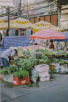 Bustling street market stall with fresh vegetables and vendors under bright umbrellas.