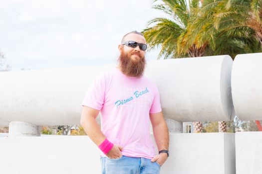 Bearded man in sunglasses posing casually outdoors with palm trees in the background.