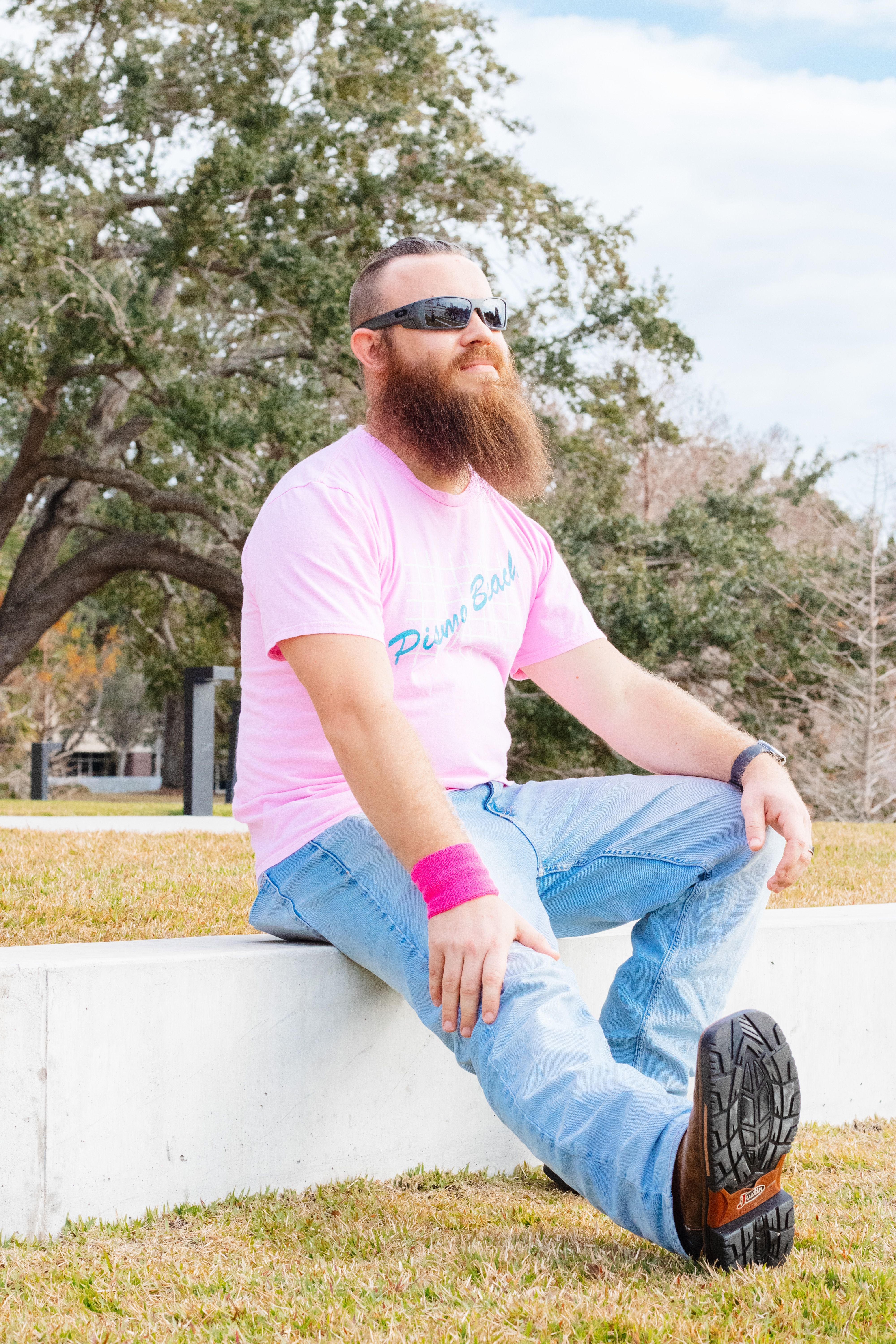 A bearded man wearing sunglasses and pink t-shirt sits joyfully on a concrete ledge outdoors.