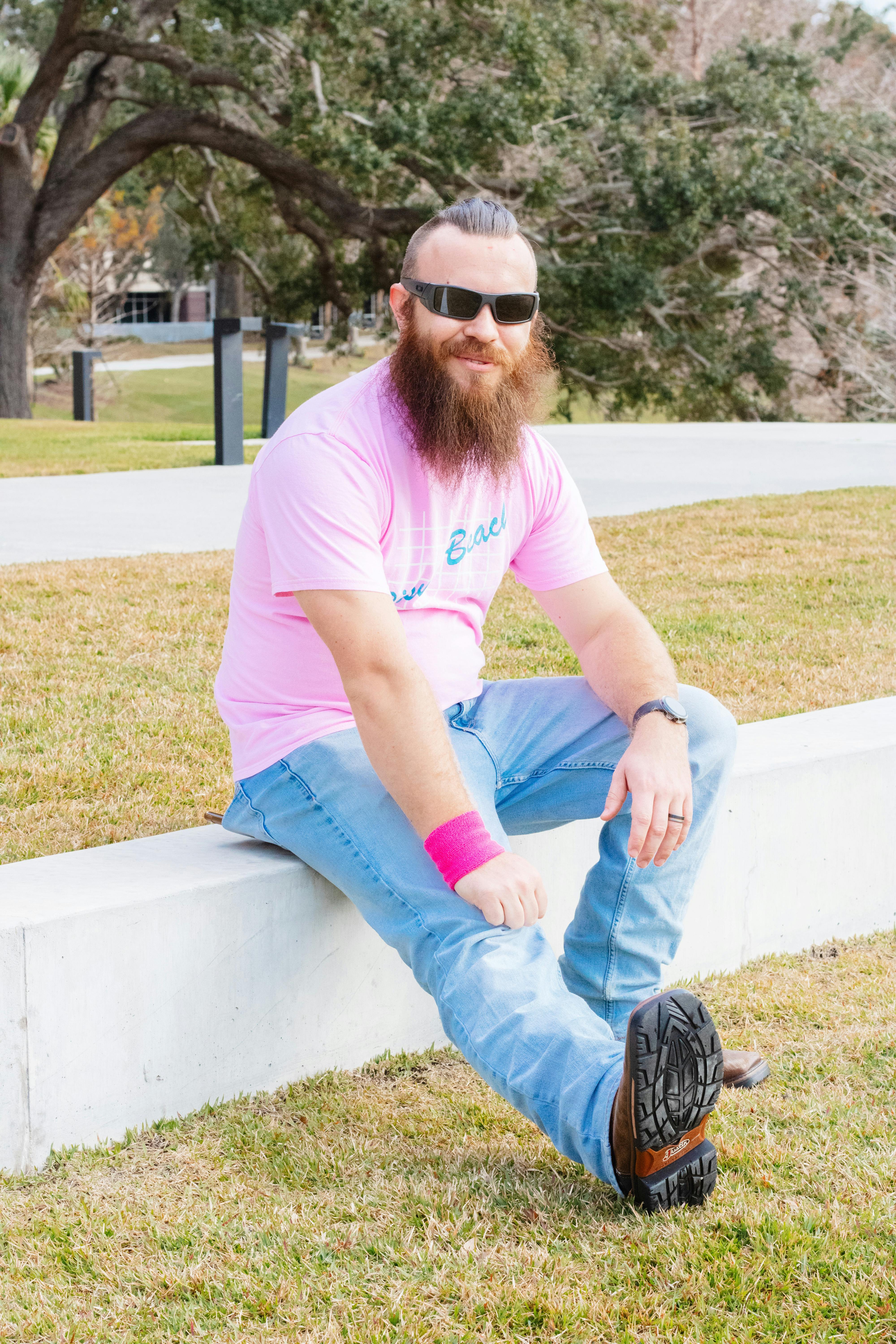 Man with beard in pink t-shirt sitting outdoors, exuding casual style.