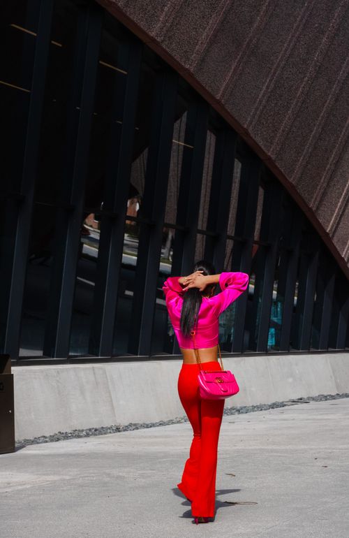 Free Fashionable woman with long hair in pink and red outfit walking outside a modern building. Stock Photo