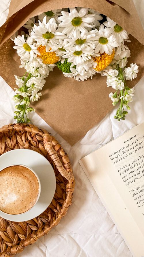 Free A cozy setup with a cup of coffee, flowers, and an open book on a woven tray. Stock Photo