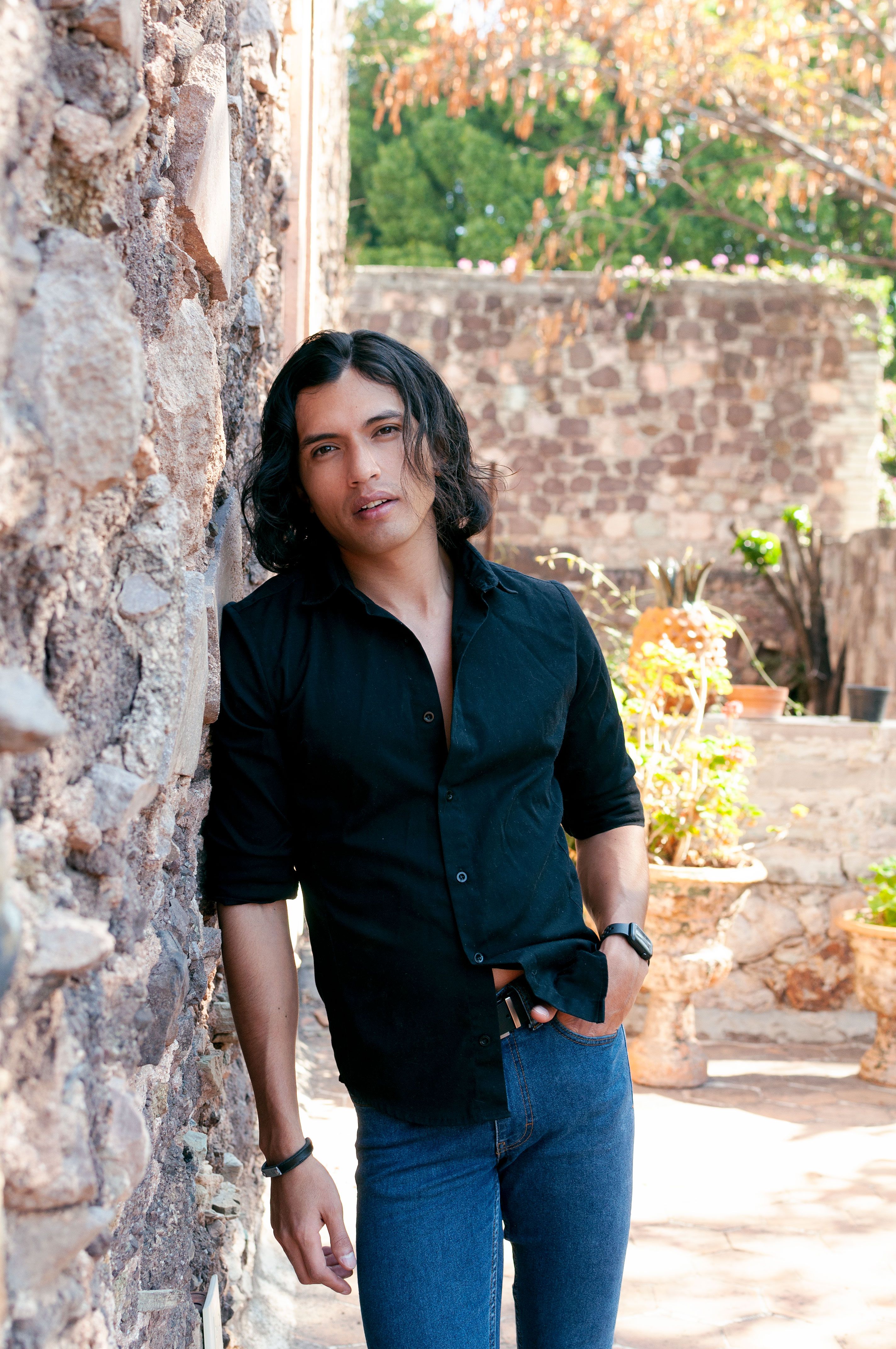 Stylish man with long hair in black shirt leaning against a rustic stone wall outdoors.