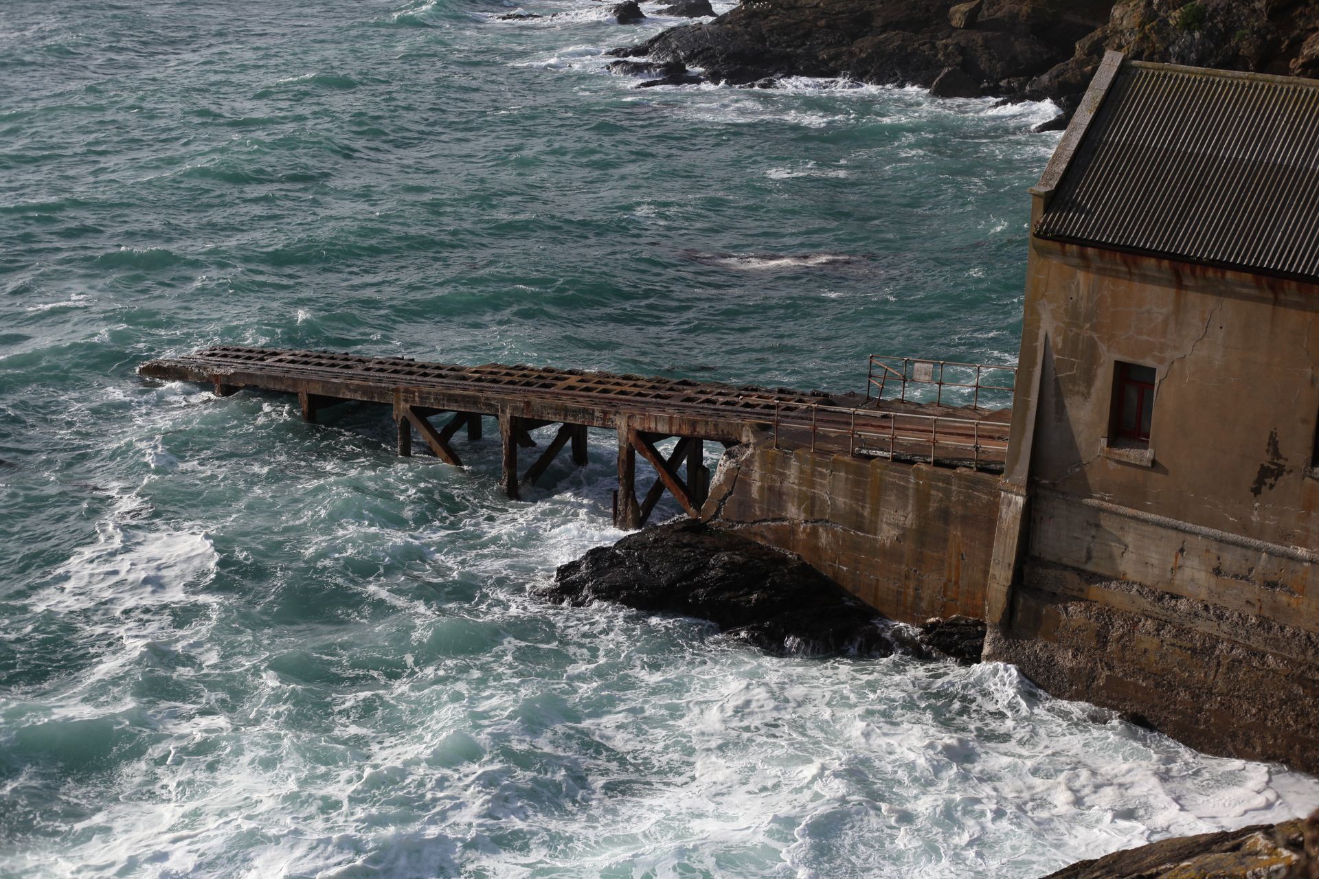 Rustic seaside building and pier at Polpeor Cove, Cornwall surrounded by splashing waves.