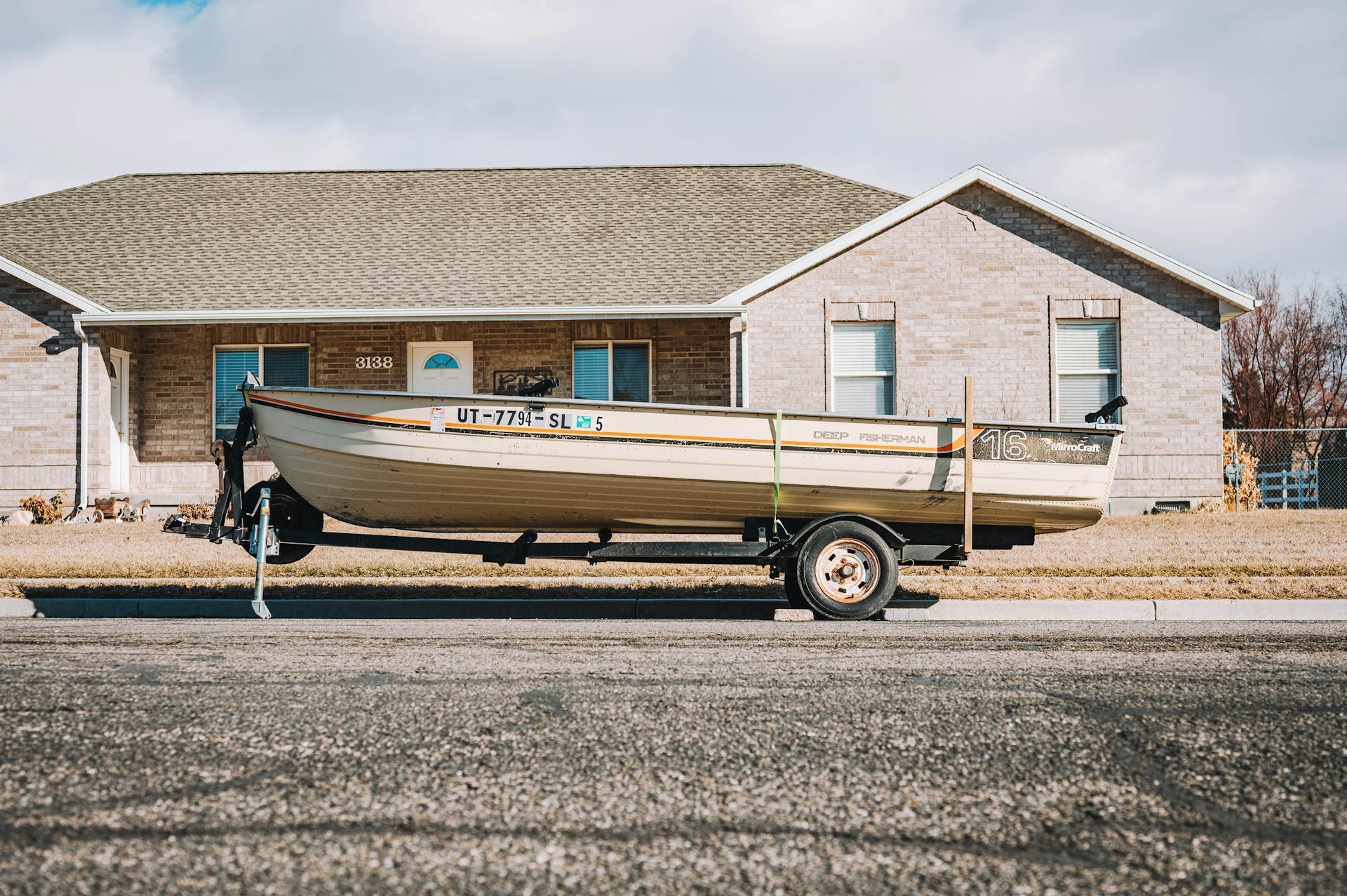 A fishing boat on a trailer parked in front of a suburban house on a clear day.