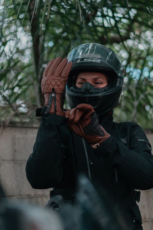Free An adult motorcyclist putting on gloves and helmet outdoors with greenery backdrop. Stock Photo