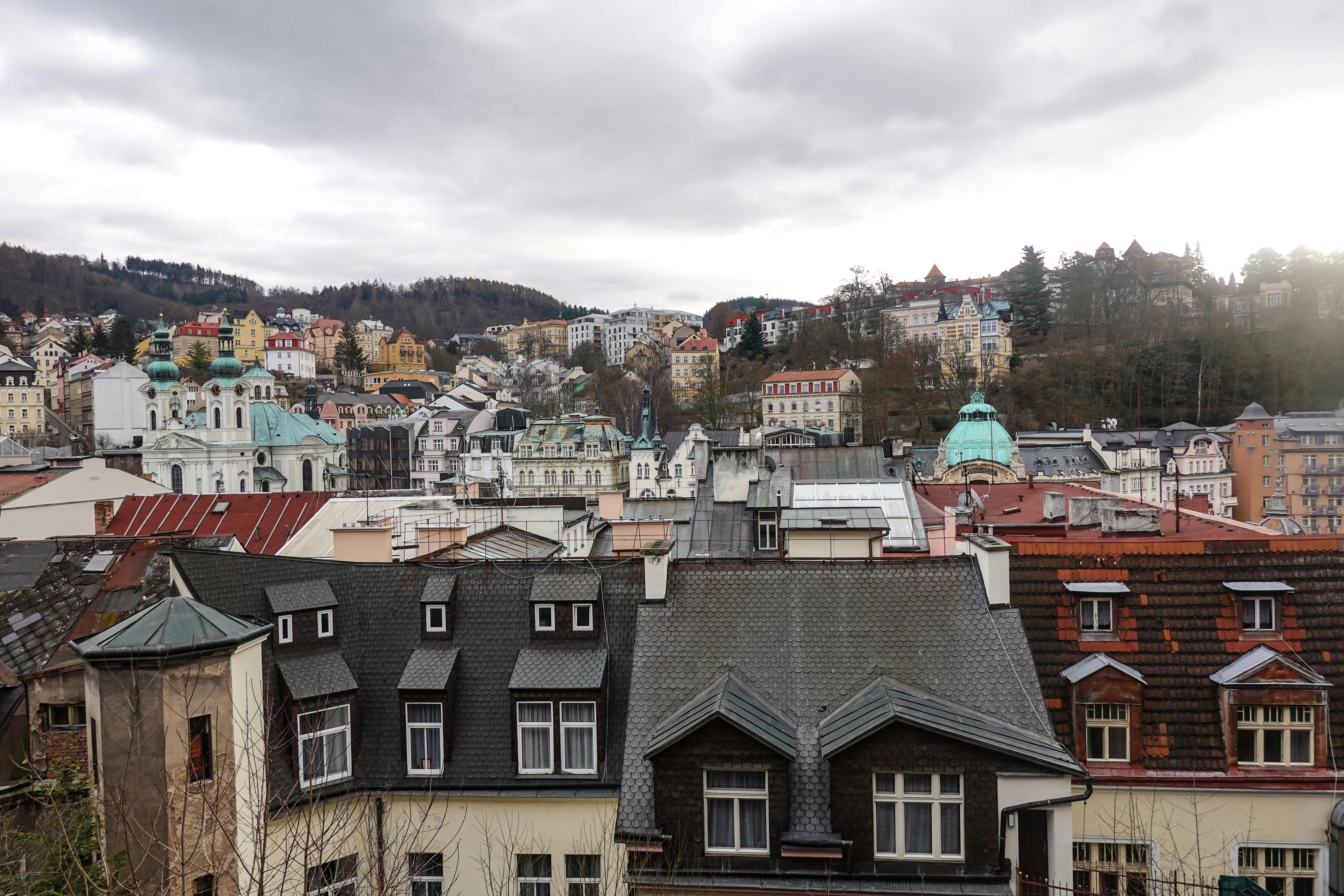 A scenic view of Karlovy Vary's historic buildings amidst lush hills on a cloudy day. - Karlovy Vary