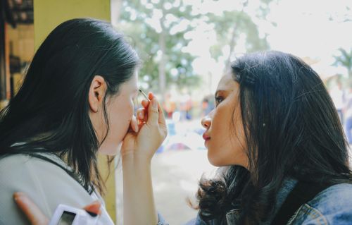 Free A young woman applies makeup to her friend outdoors, enjoying a sunny day. Stock Photo
