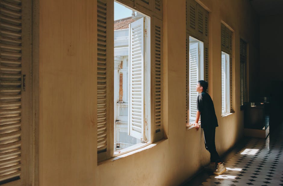A man in a hallway looking out through open wooden shutters, bathed in sunlight.