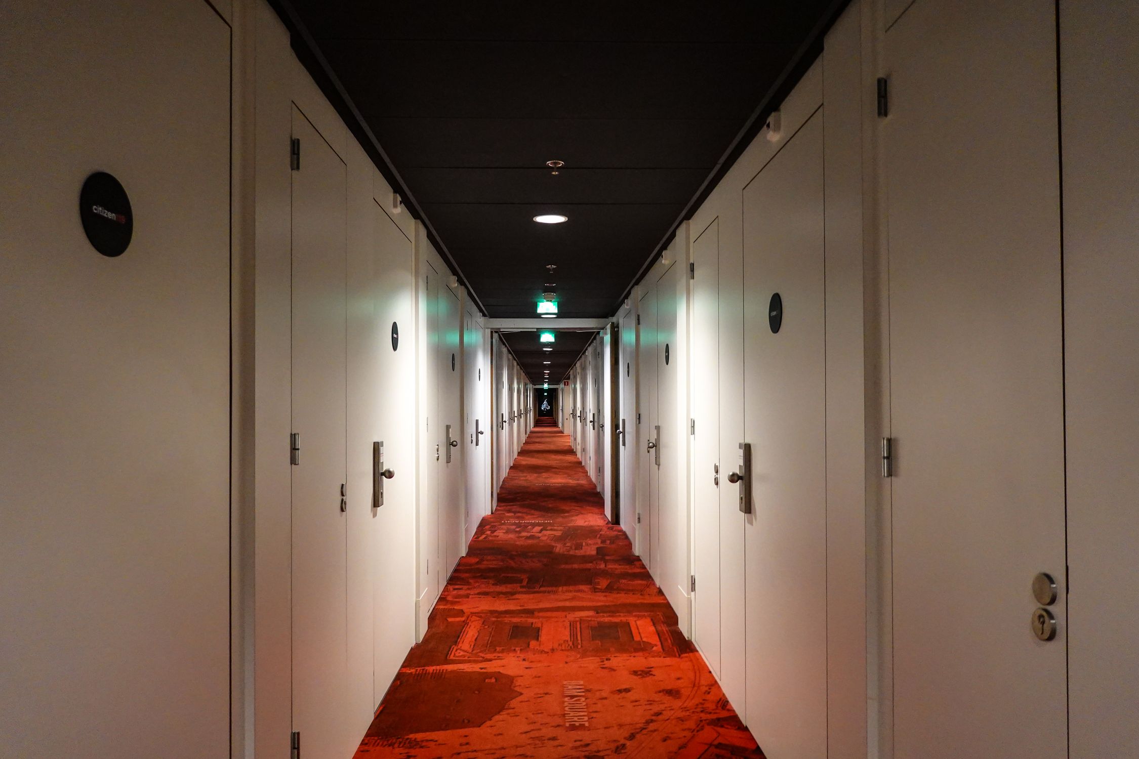 Long, symmetrical hotel hallway with red carpet and closed white doors under warm lighting.