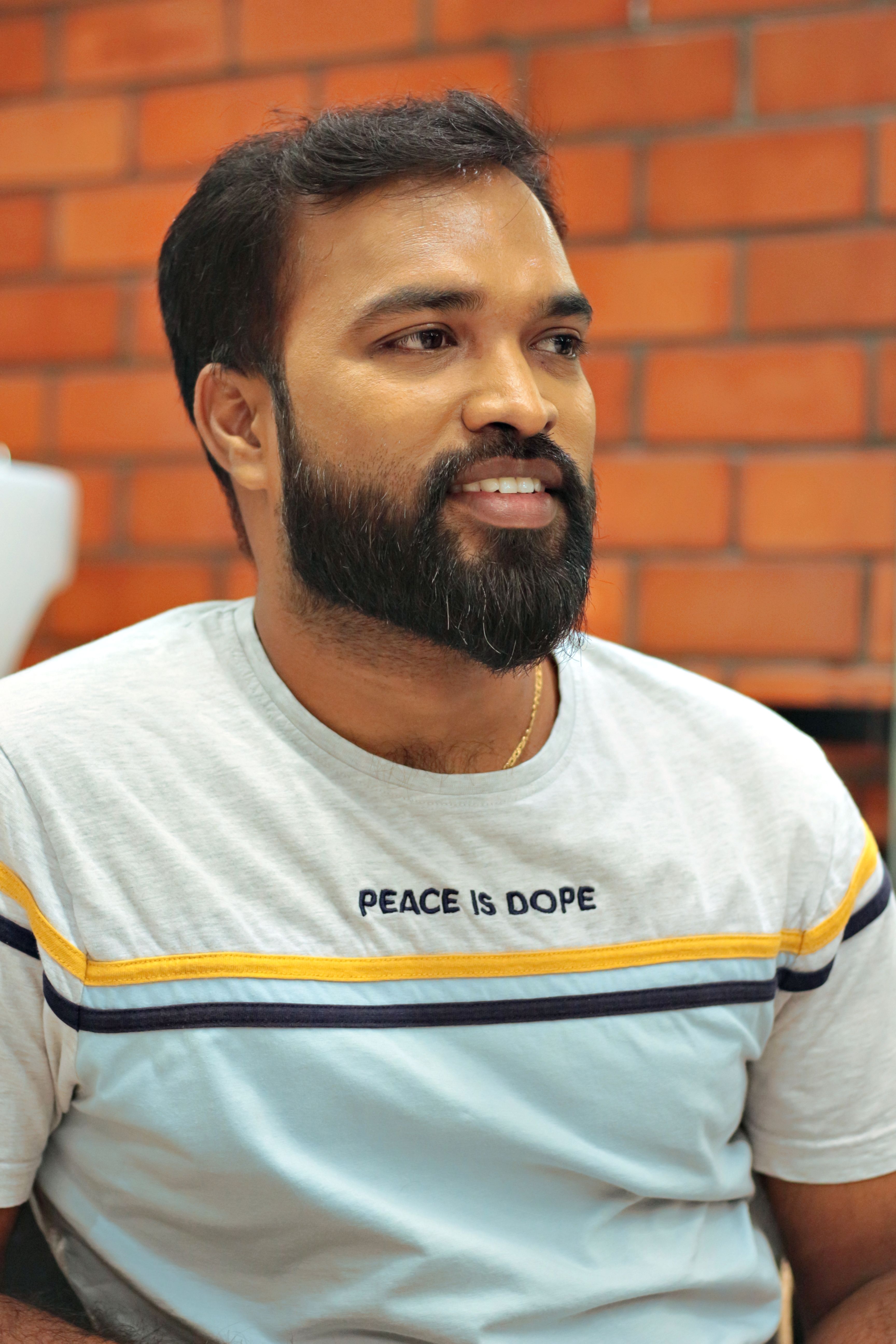 Portrait of a smiling man with a beard wearing a casual 'Peace is Dope' t-shirt against a brick wall.