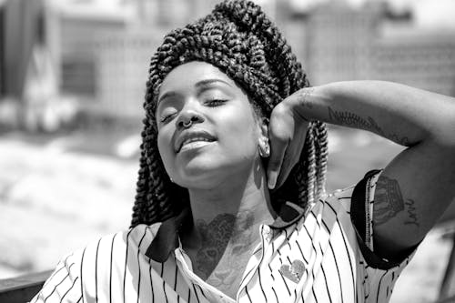 Black and White Photo of a Young Woman with Braided Hair and Tattoos Standing with Eyes Closed in Sunlight 