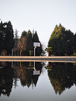 Basketball court with hoop reflecting on calm water surface under clear sky.
