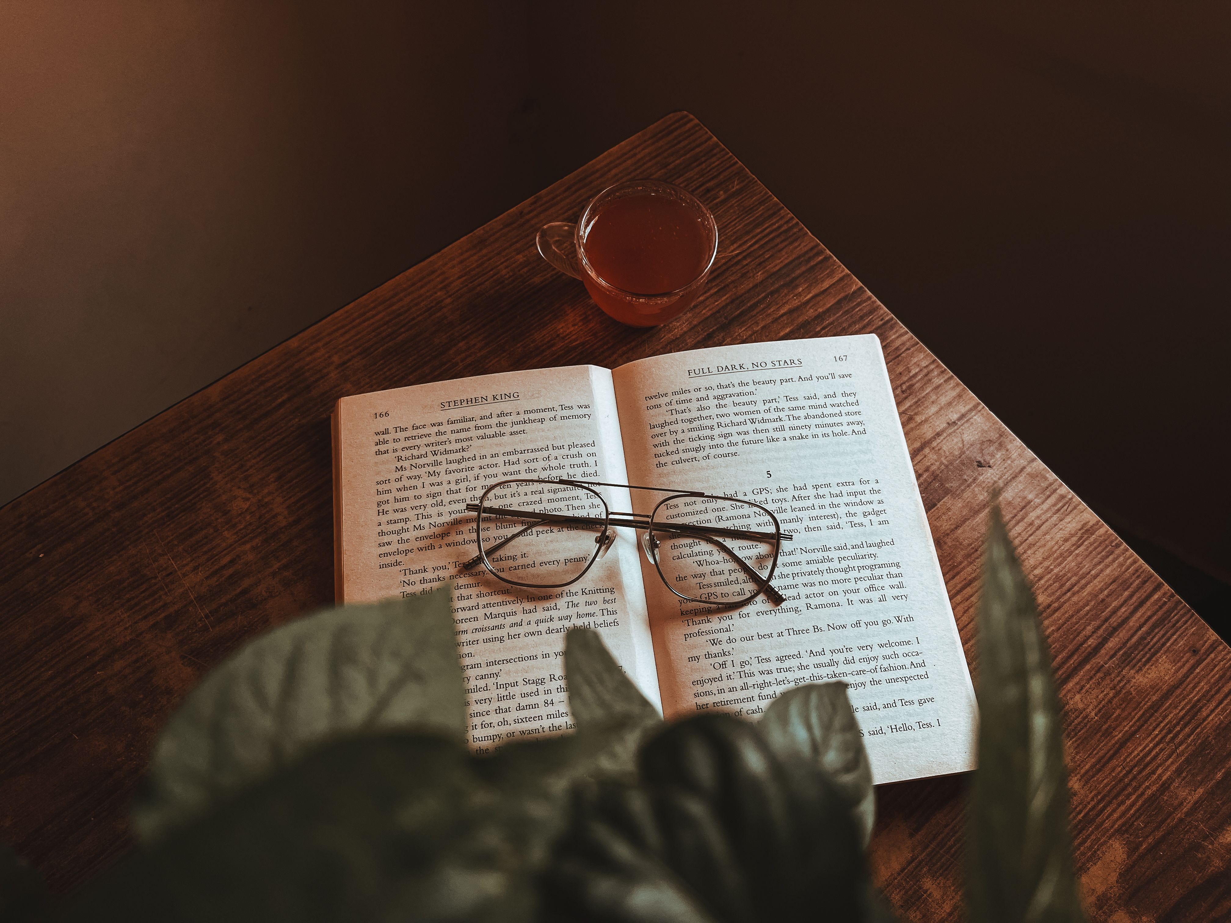 Open book with glasses and tea on wooden table, creating a serene reading atmosphere.