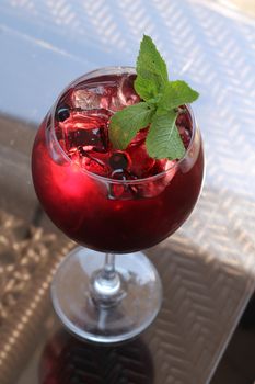 A vibrant red cocktail with mint leaves, served in a glass with ice cubes on a reflective table.