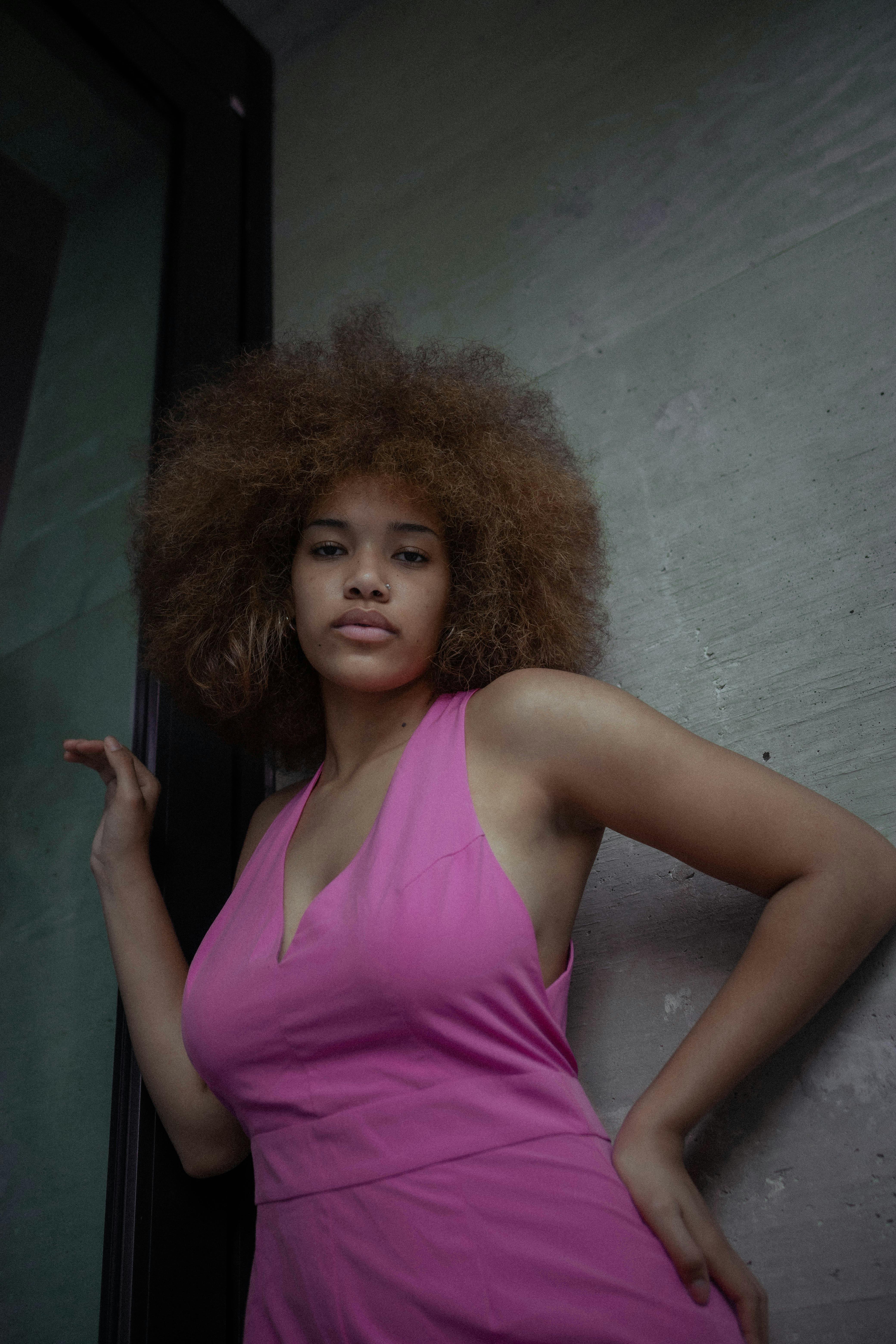 Stylish portrait of a woman with afro hair in a pink dress, leaning casually indoors.