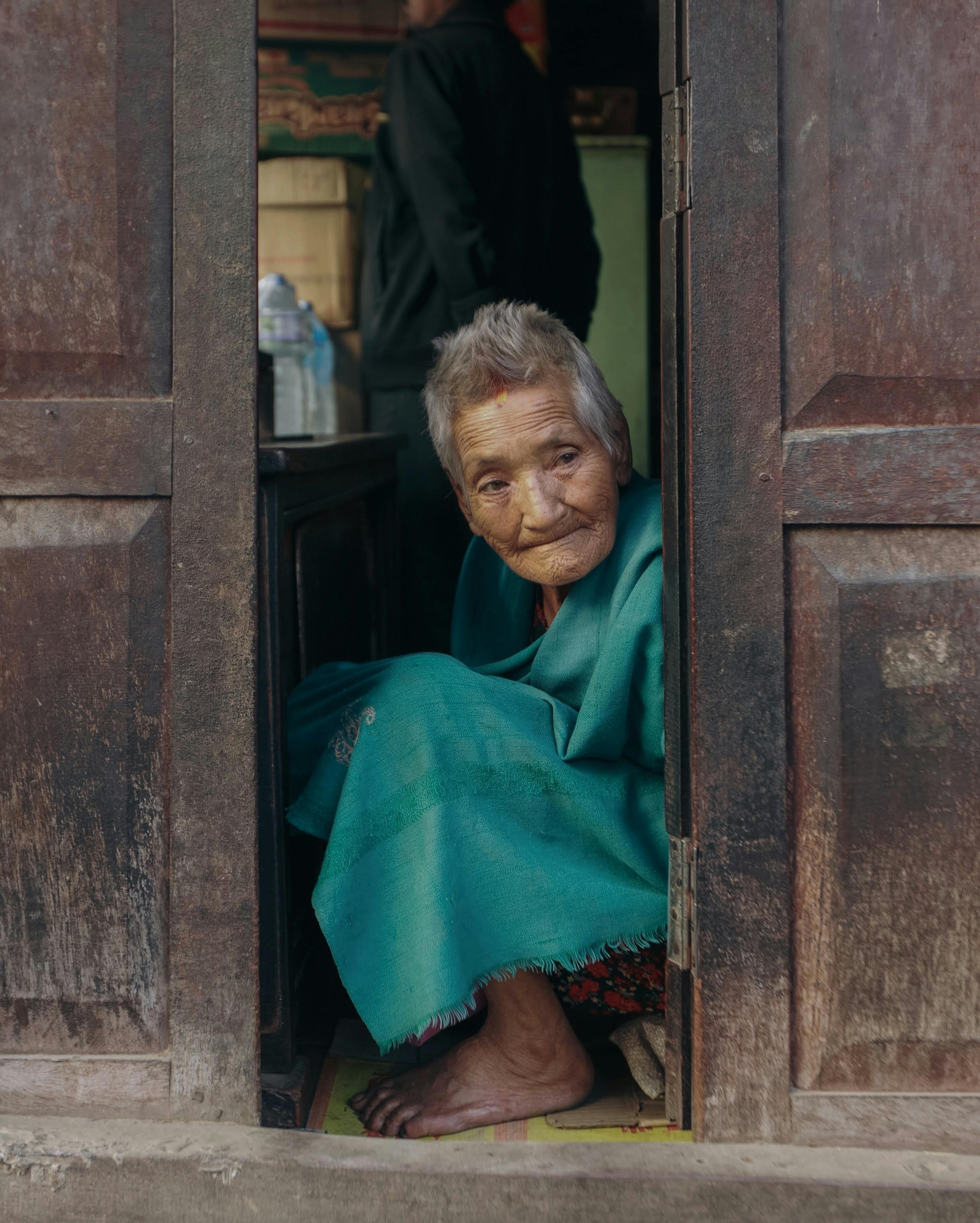 A senior woman wrapped in a turquoise blanket sits and peers through a wooden doorway.