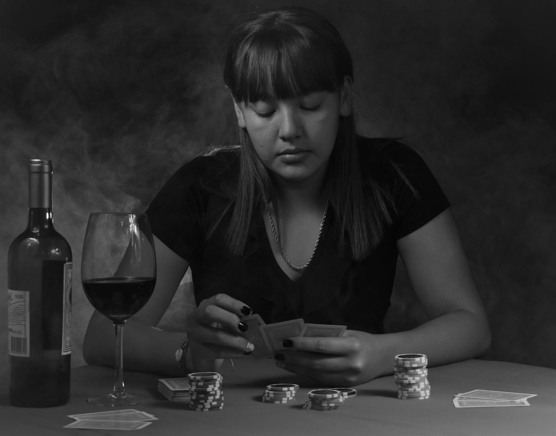 Woman focused on poker game, surrounded by wine and chips in a black and white setting.