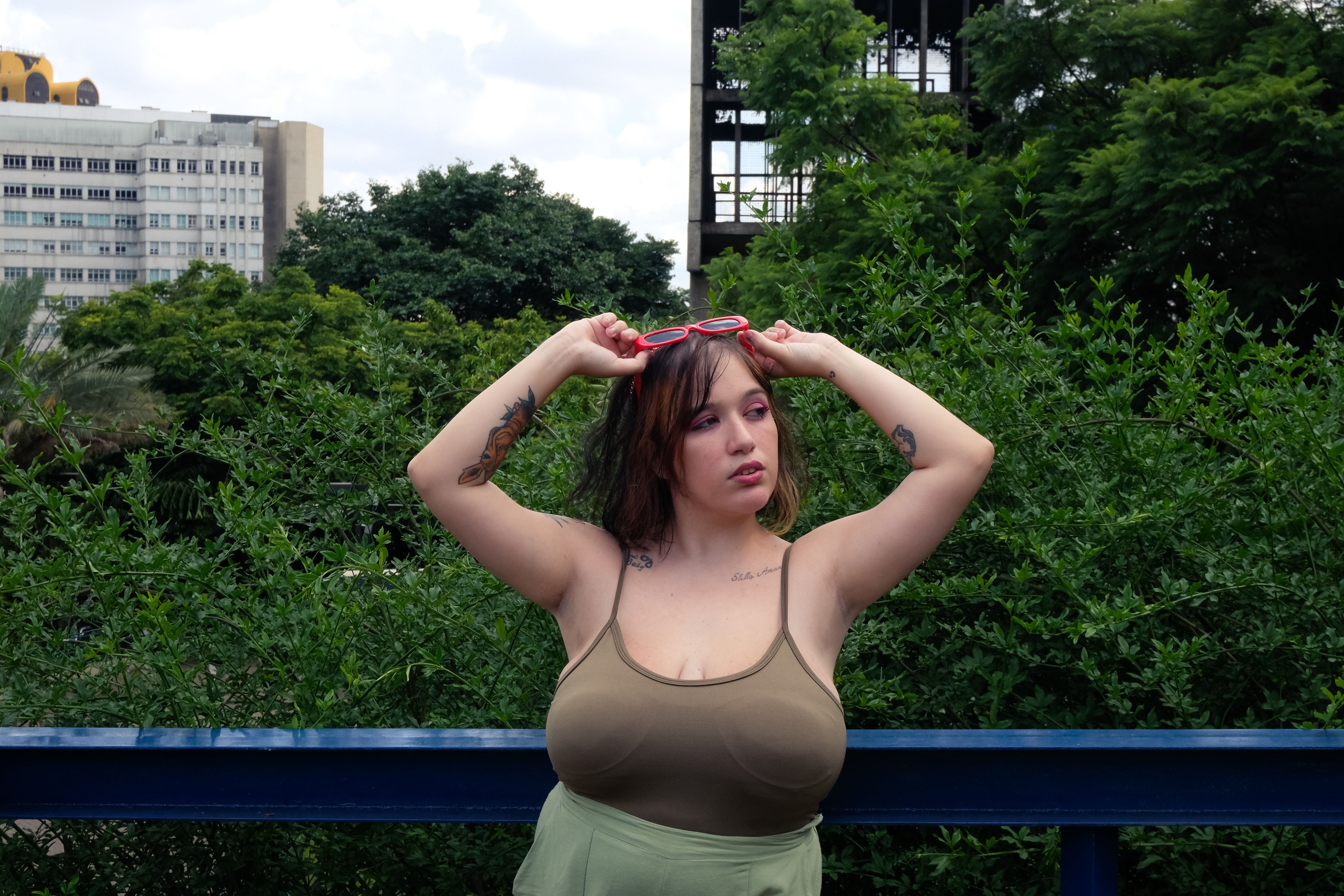 Young woman adjusting red sunglasses outside against a verdant backdrop in São Paulo.