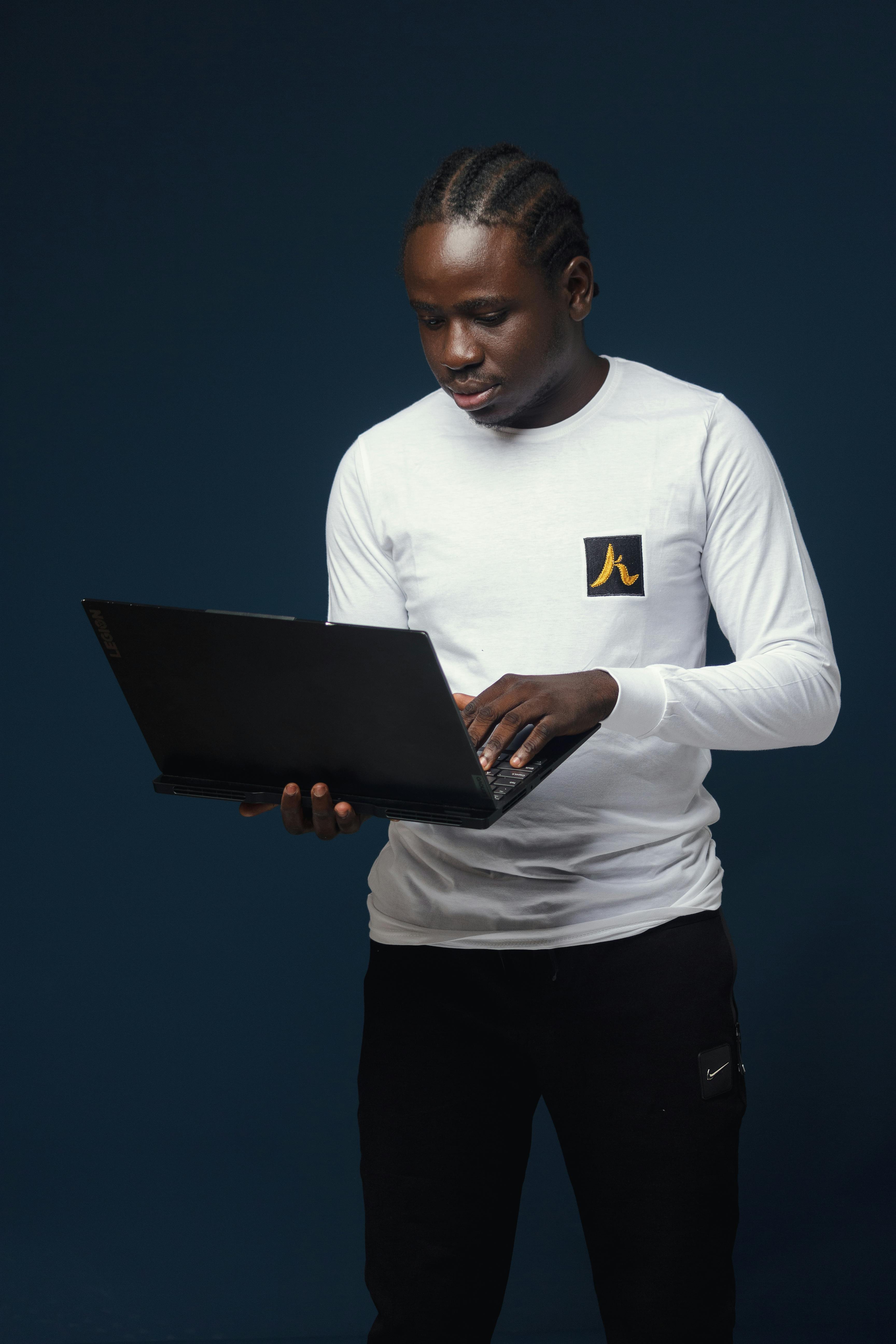 Black man in a white shirt stands against a blue background using a laptop.