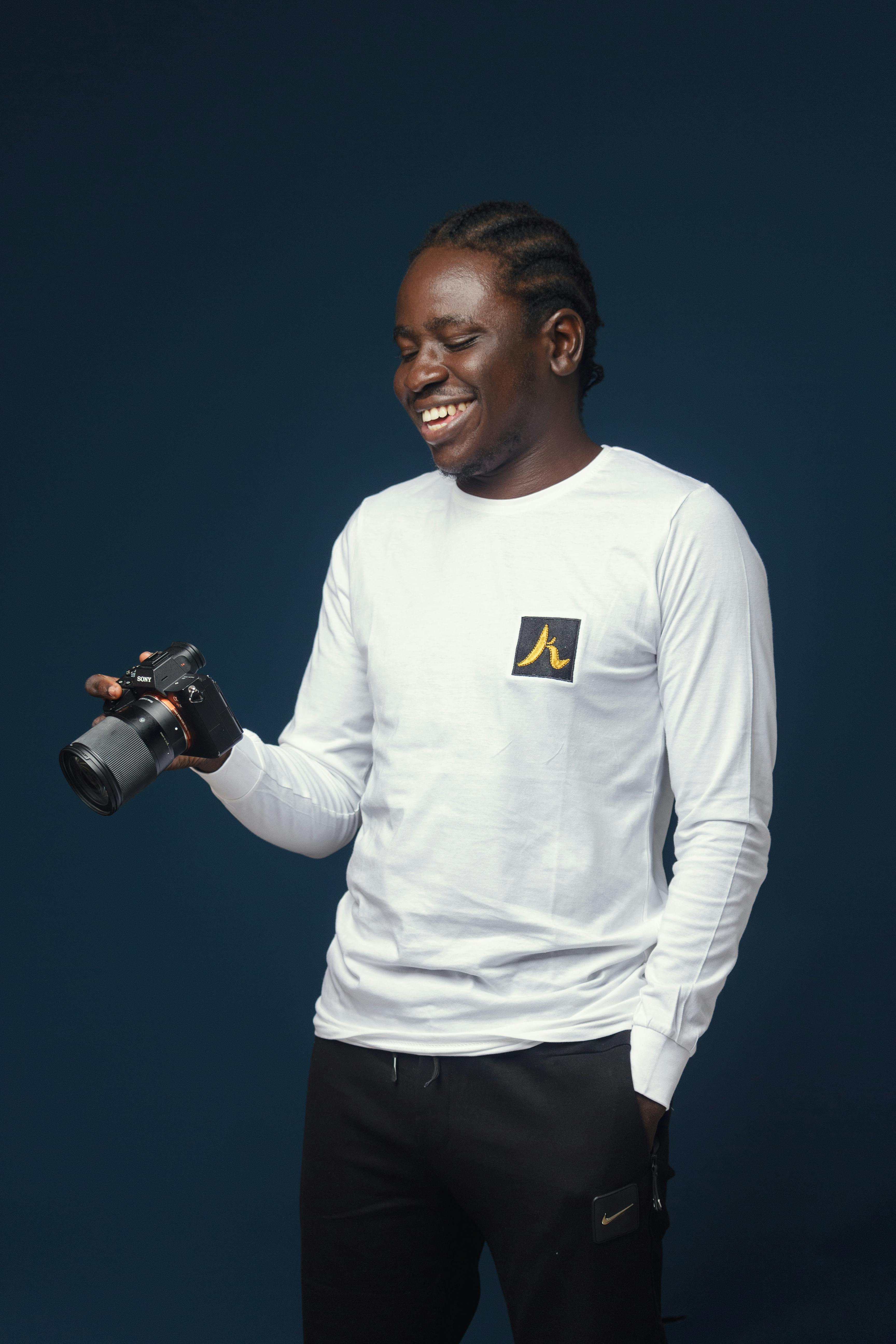 Portrait of a smiling man holding a camera, wearing a white shirt against a dark background.
