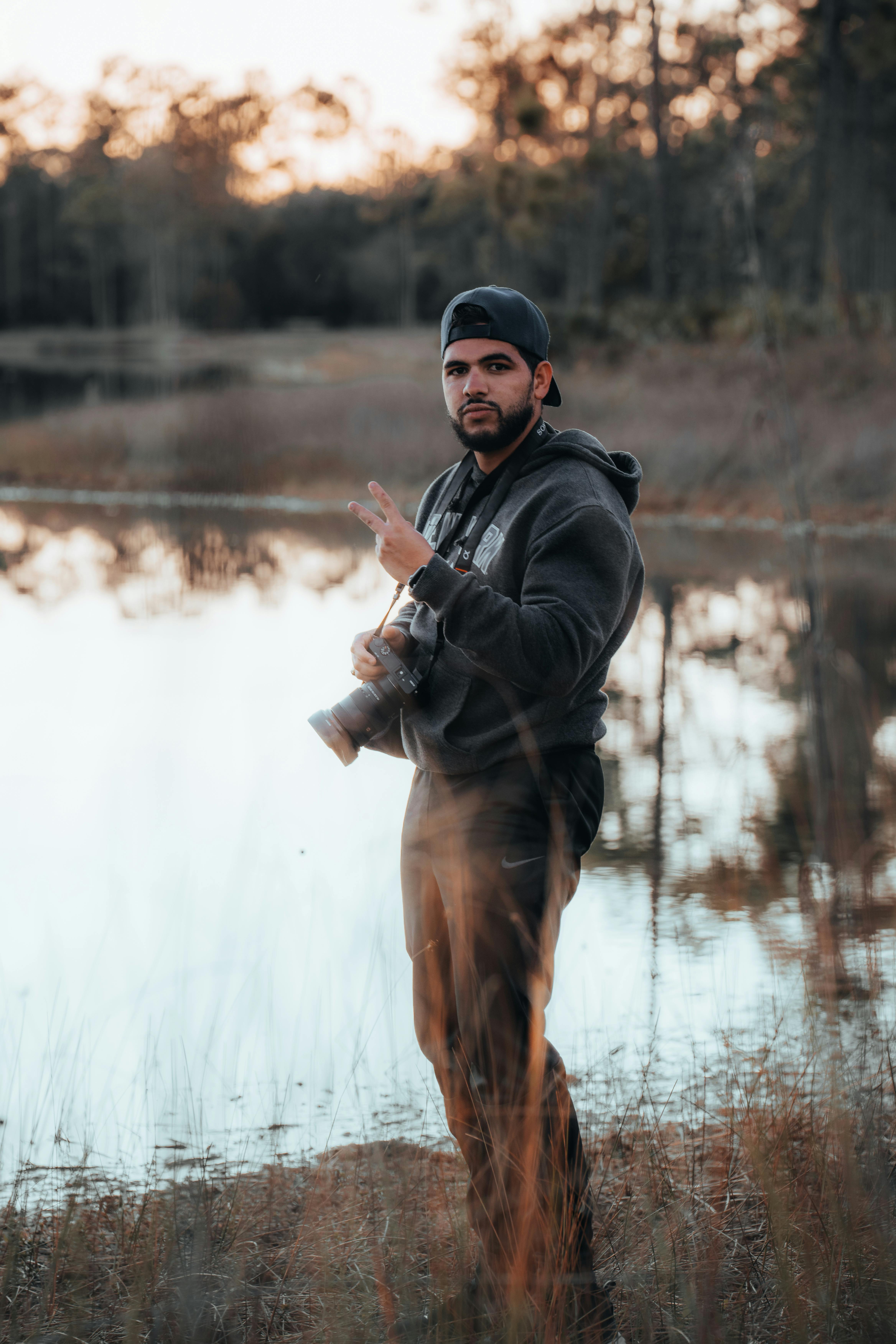A man with a camera poses by a tranquil lake, flashing a peace sign outdoors.