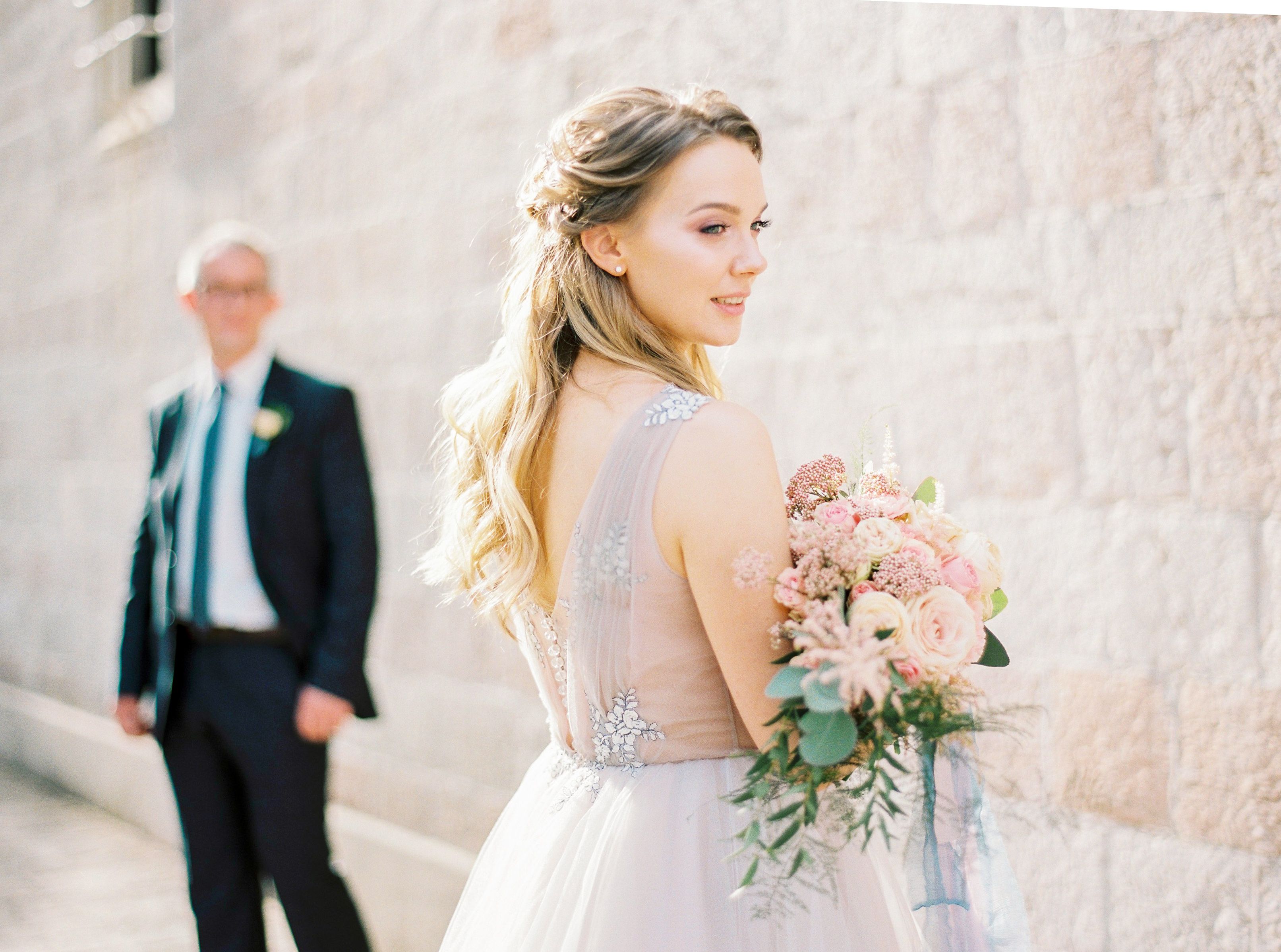 A beautiful bride holding a bouquet, posing near a stone wall with a groom in the background.