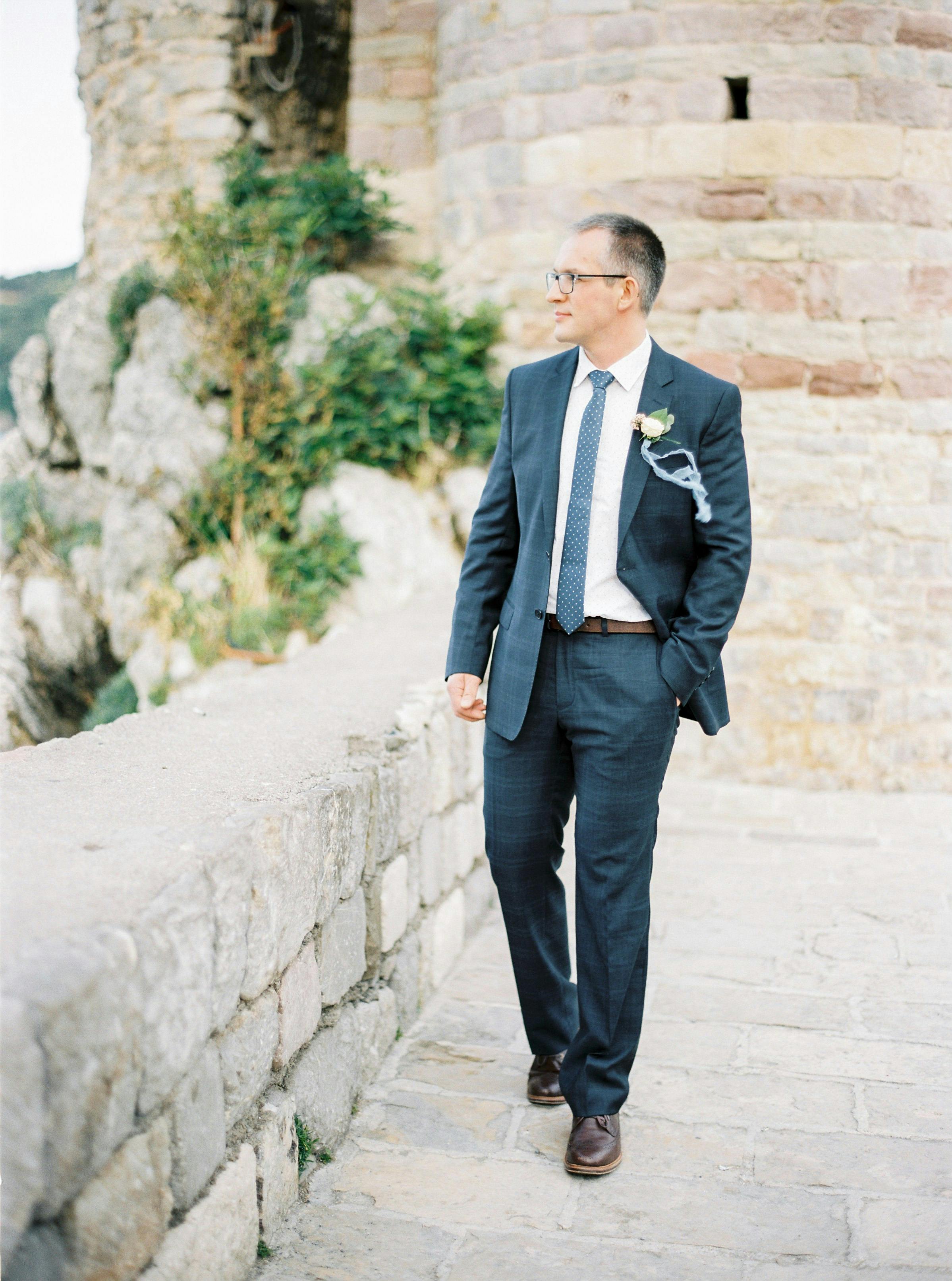 Groom in a formal suit walking along the stone path in a historic town, showcasing elegance and style.