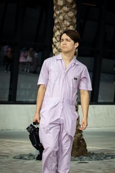 A man in a purple jumpsuit holding a camera walks outdoors by a palm tree.