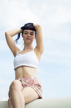 Young woman posing in casual summer outfit against a clear sky, exuding a relaxed and playful vibe.
