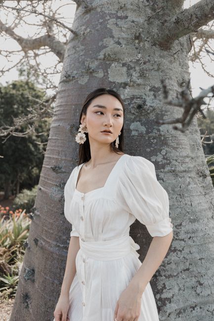 Elegant woman poses in white dress with flower earrings beside a large tree trunk outdoors.