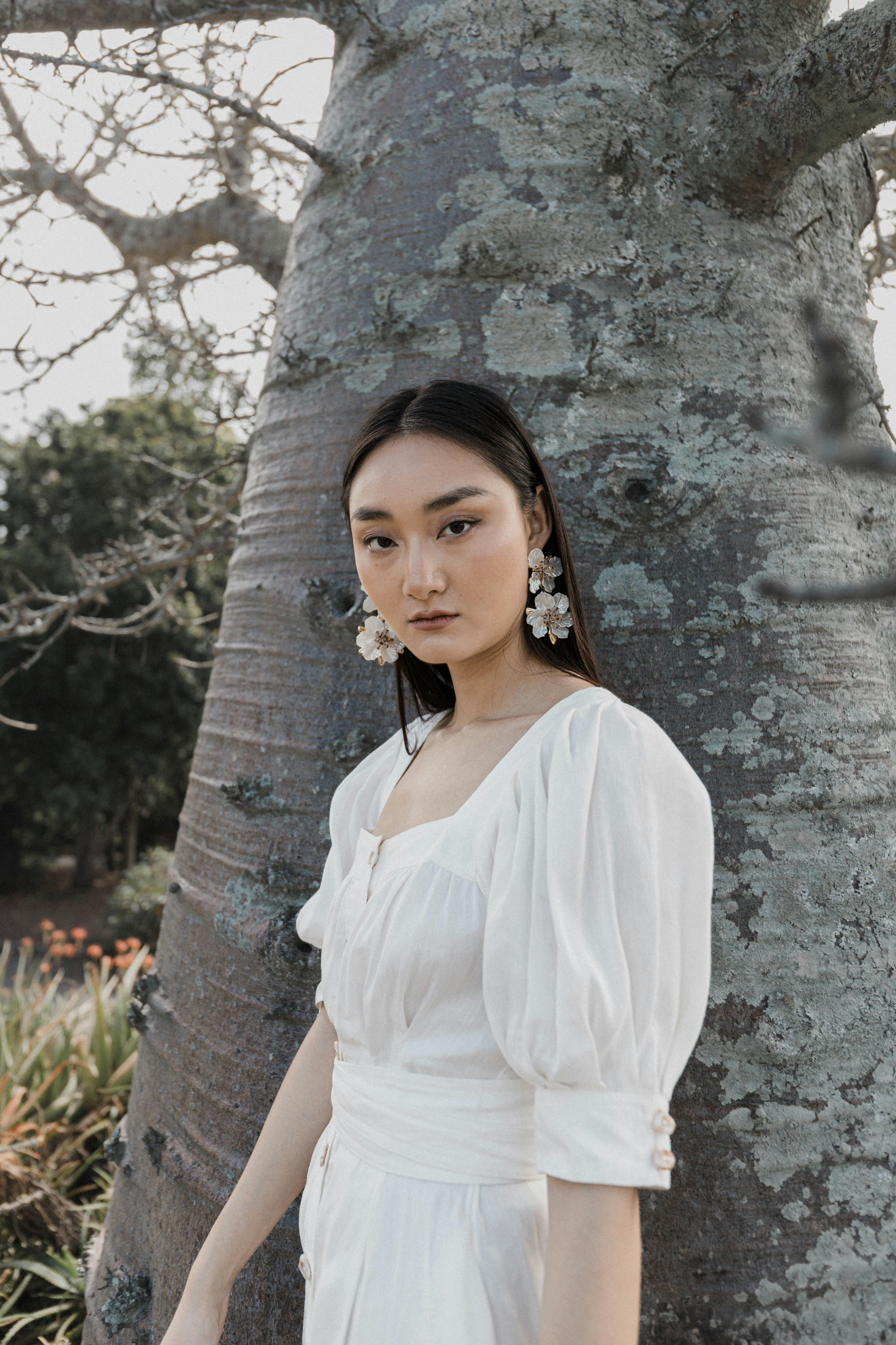 A portrait of an elegant woman in a white dress leaning against a tree outdoors, exuding grace and style.