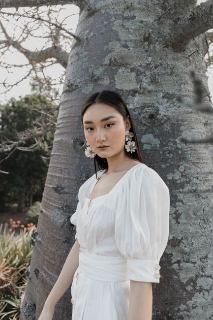 A portrait of an elegant woman in a white dress leaning against a tree outdoors, exuding grace and style.
