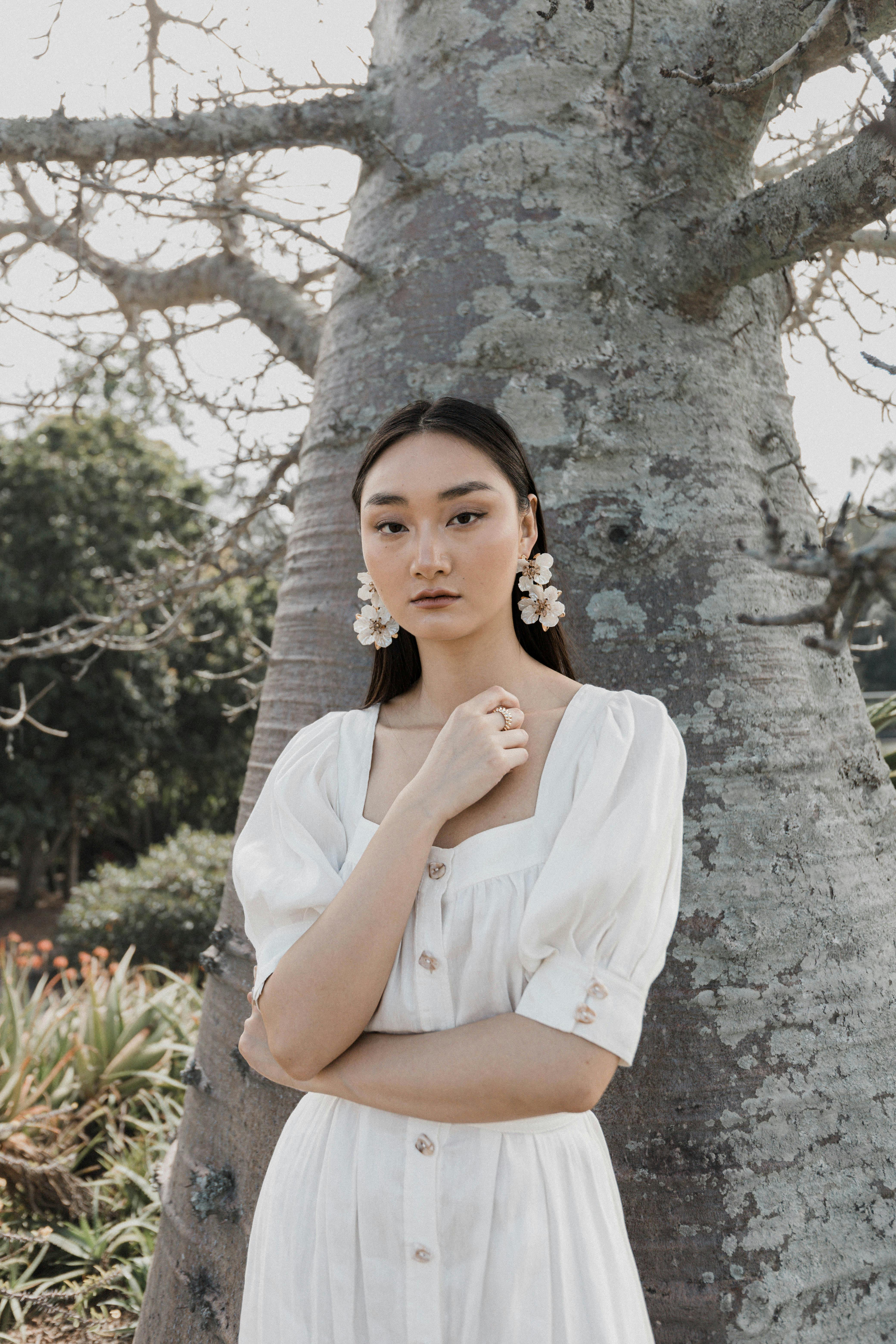 Stylish Asian woman in white dress with floral earrings standing near a tree outdoors.