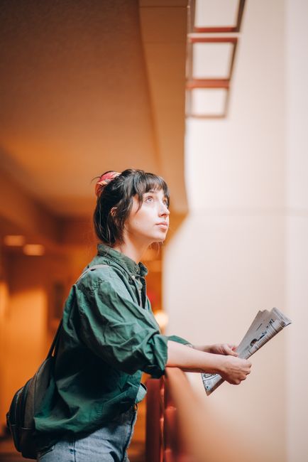 A thoughtful young woman in a green shirt holds a newspaper indoors, looking upward.