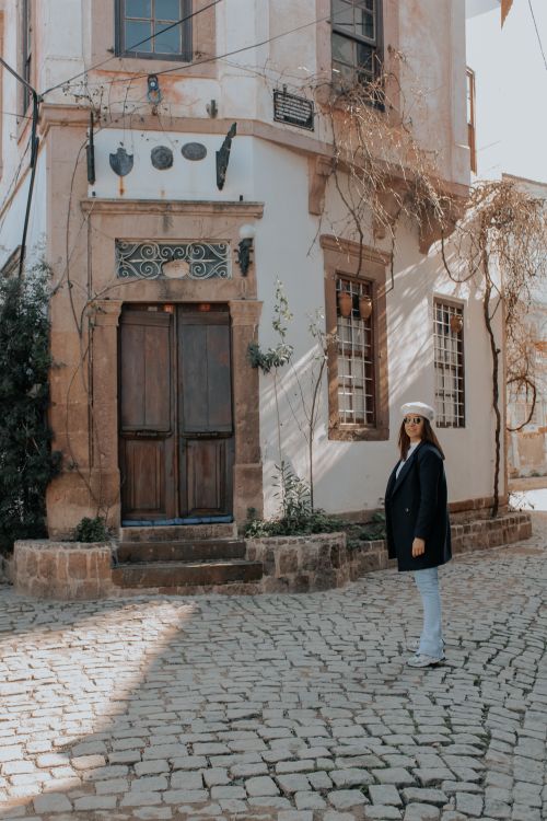 Free Woman in coat stands on cobblestone street beside a rustic wooden door in a quaint town. Stock Photo