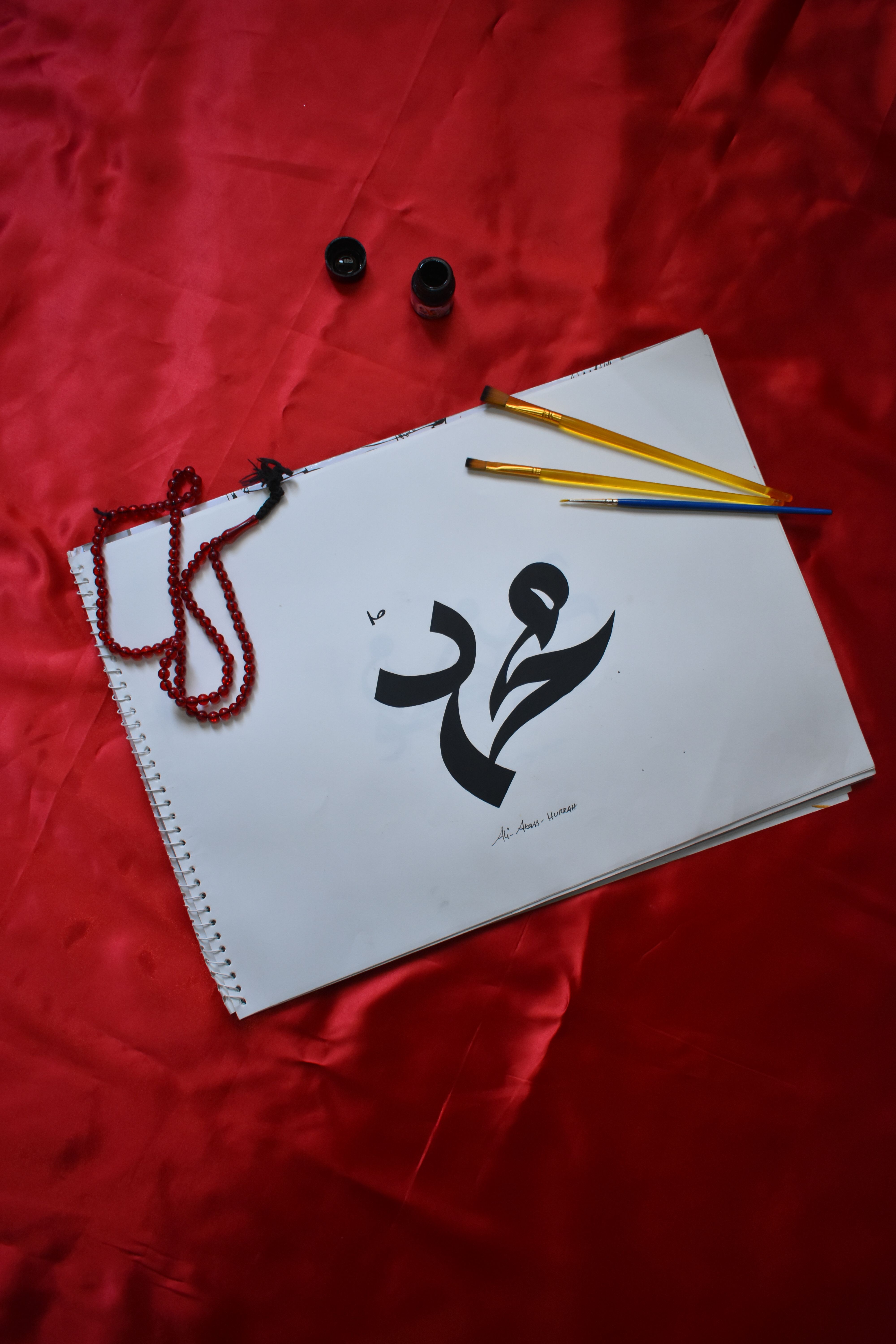 Close-up of Arabic calligraphy on sketchpad with brushes and beads on red fabric.