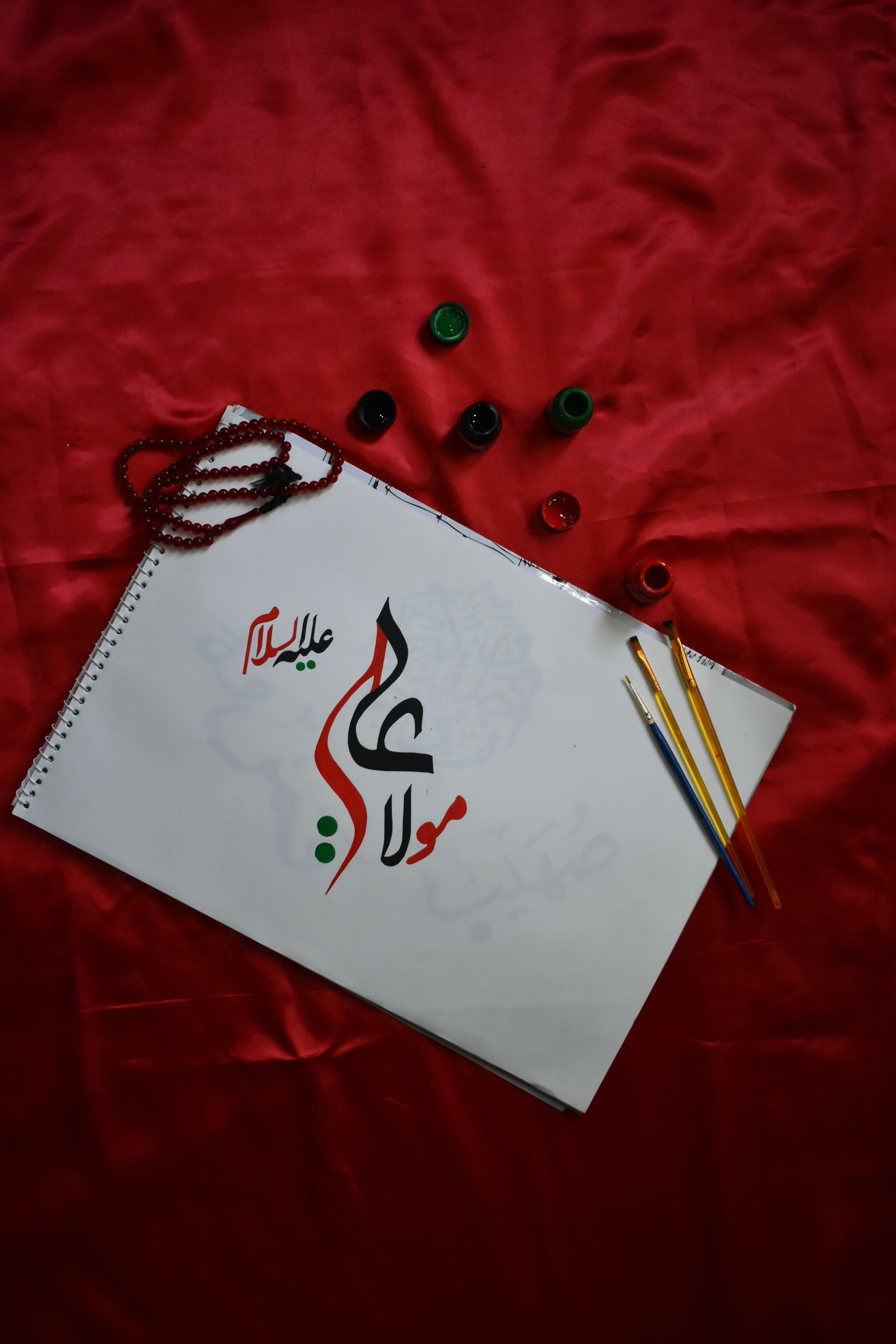 Arabic calligraphy art with paintbrushes and beads on a red fabric background.