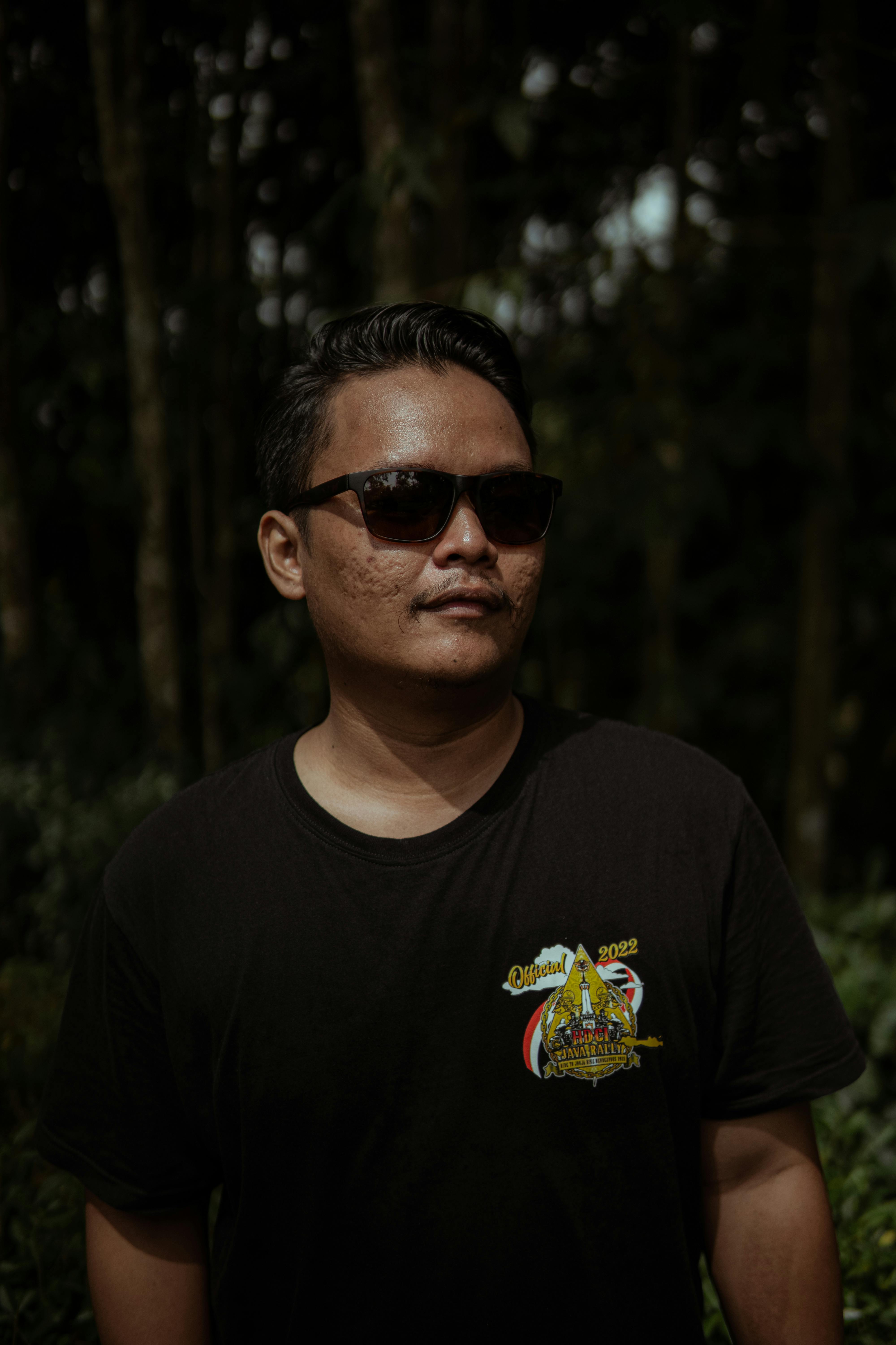 Portrait of a man in sunglasses and a black t-shirt standing outdoors with a forest background.