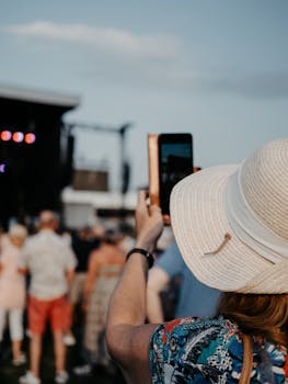 A woman in a sun hat takes photos at an outdoor music festival with her smartphone.