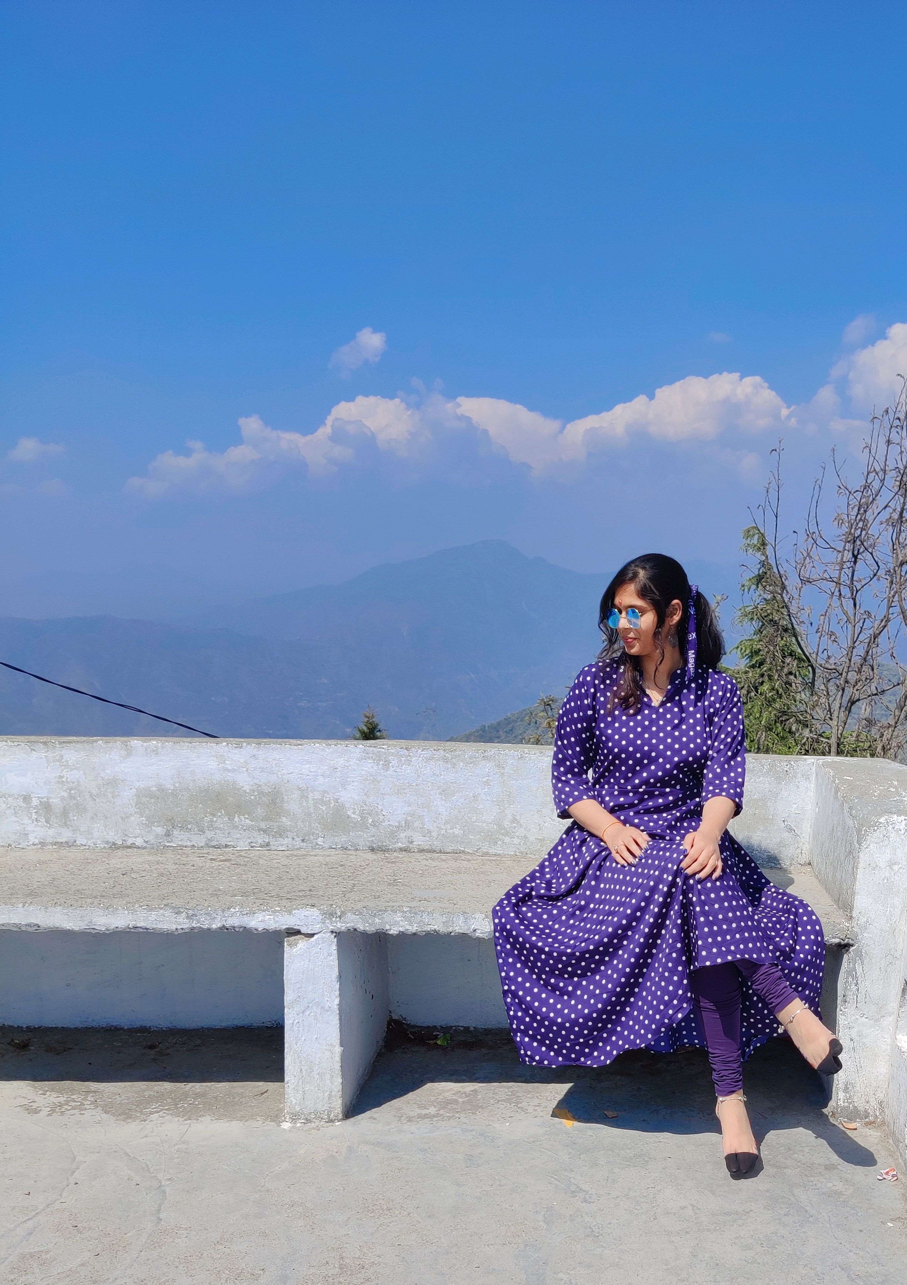 A woman in a polka dot dress sits on a bench against a mountain backdrop, enjoying a sunny day.