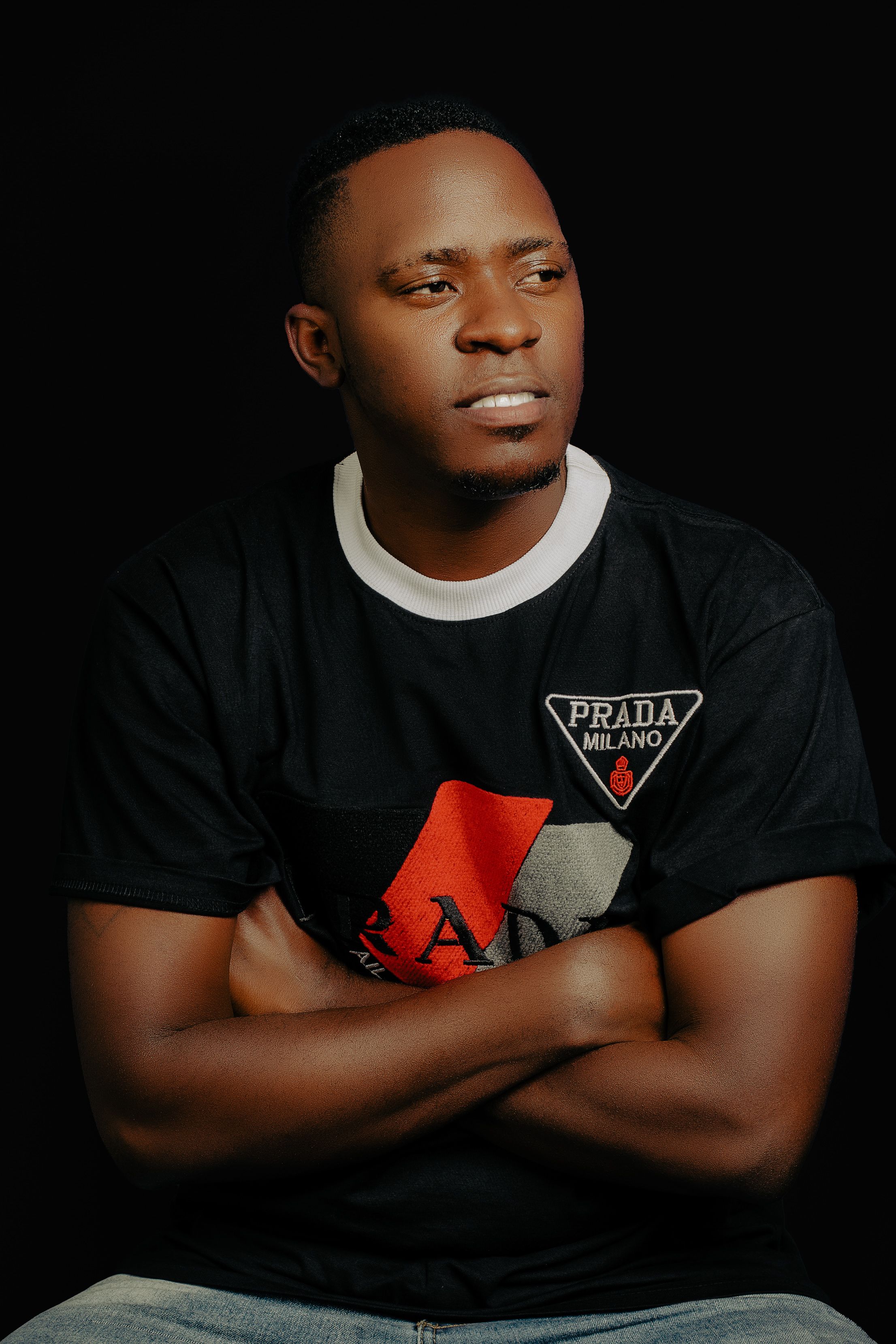 A confident man sitting with arms crossed, wearing a branded black t-shirt against a dark background.