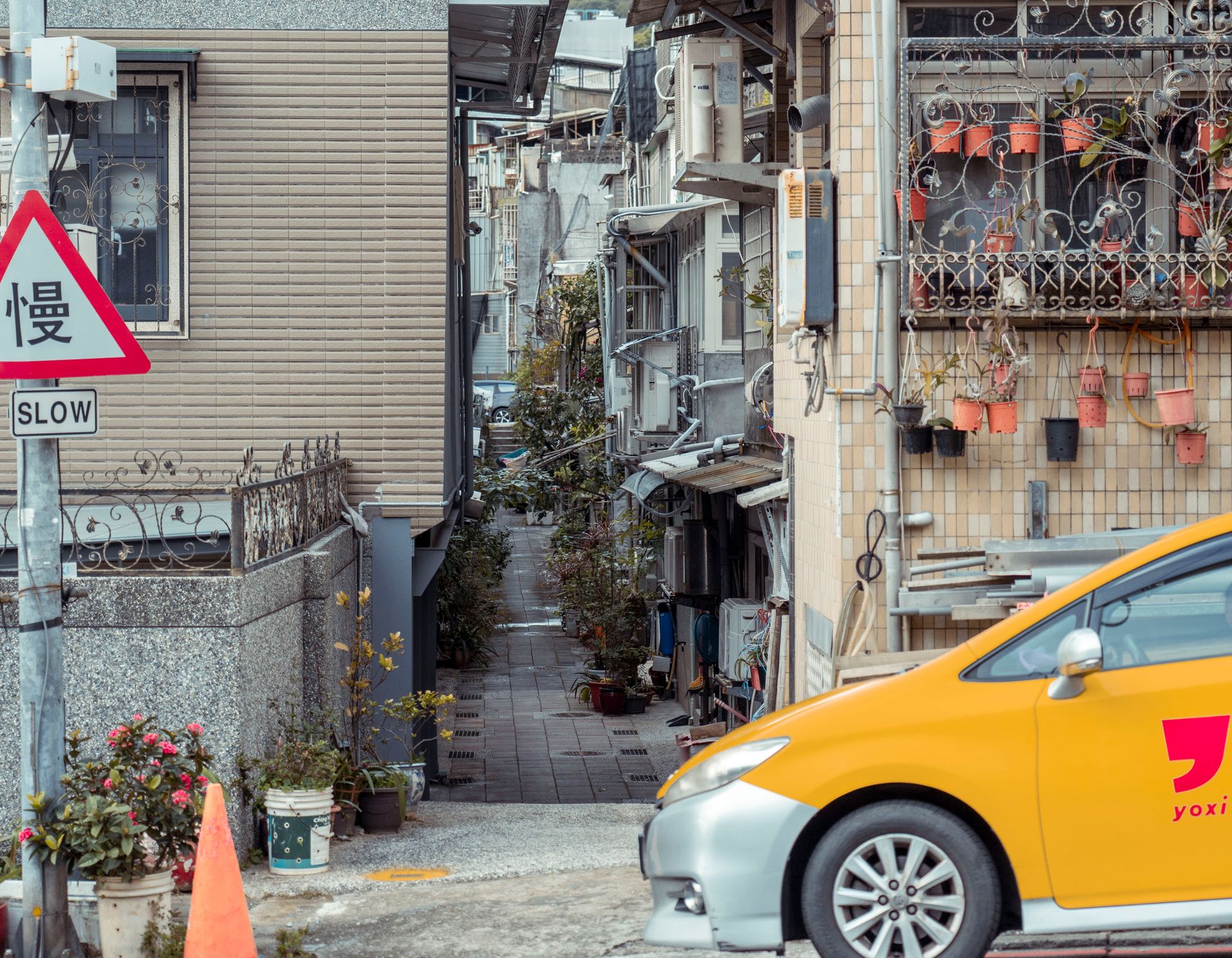 A narrow alleyway flanked by residential buildings, featuring a slow sign and a parked taxi in an urban setting.