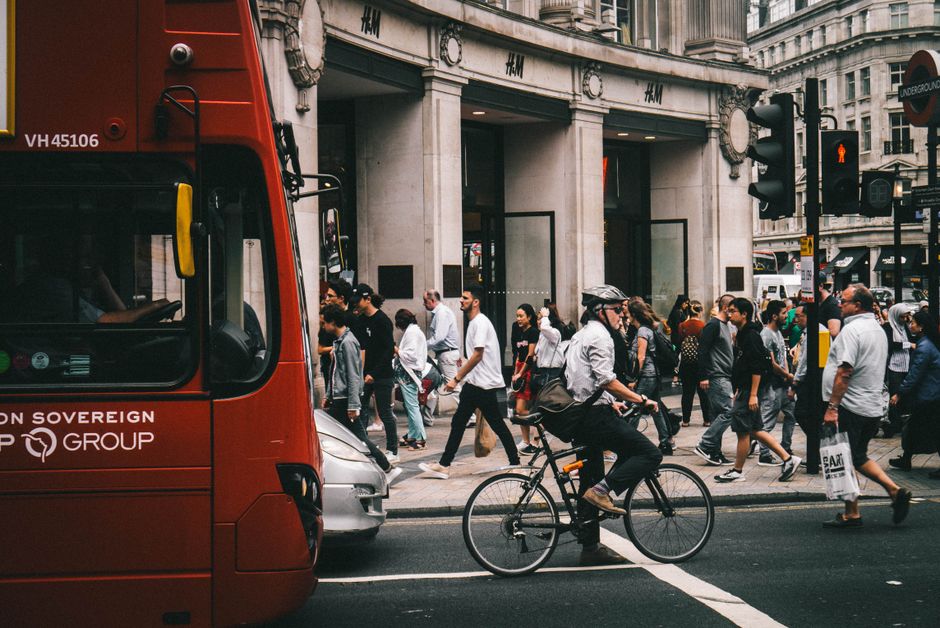 Crowd crossing London street with cyclist and iconic red bus, showcasing urban life.