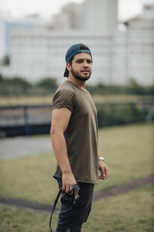 Free Man wearing a baseball cap holds a camera outdoors, showcasing a casual lifestyle. Stock Photo