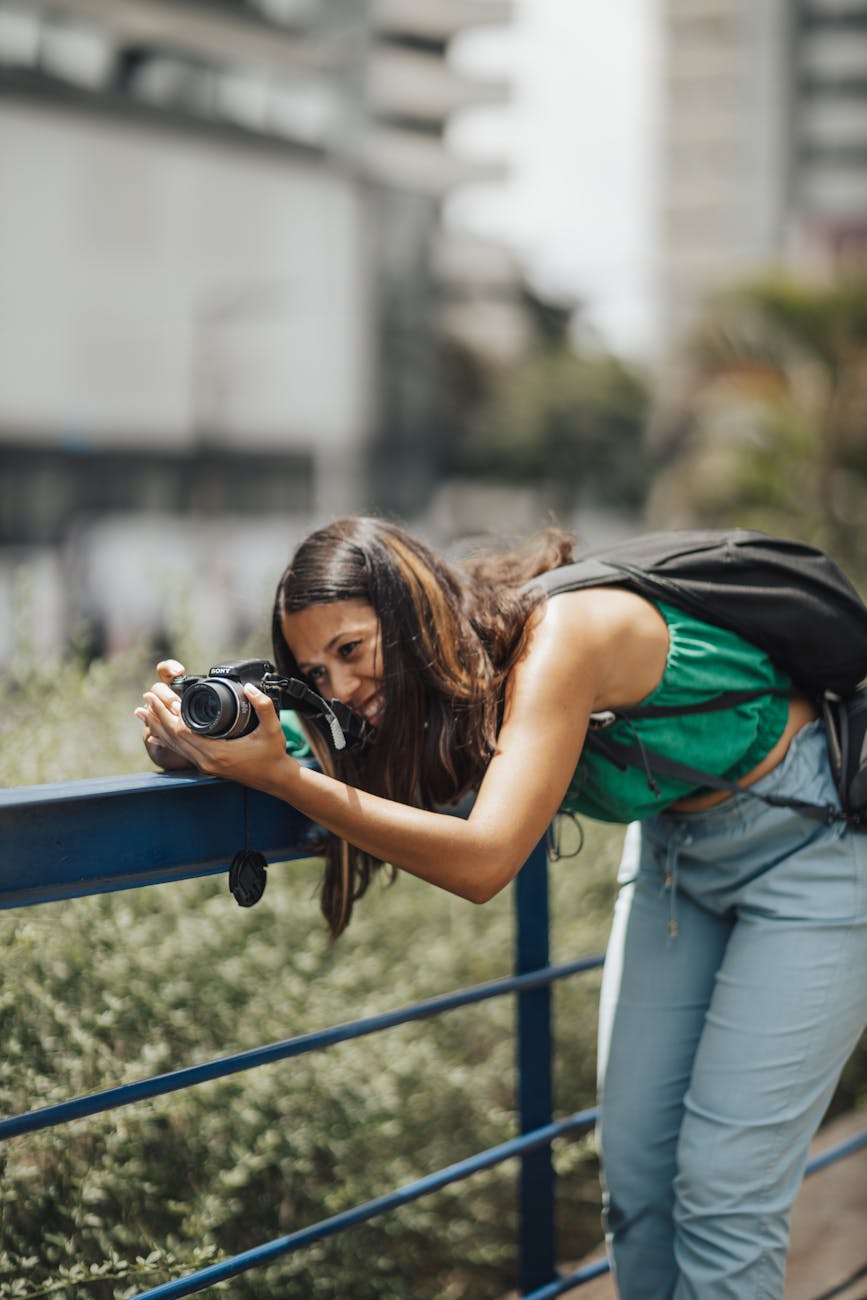 Young woman photographing cityscape with DSLR, leaning over a blue railing outdoors.